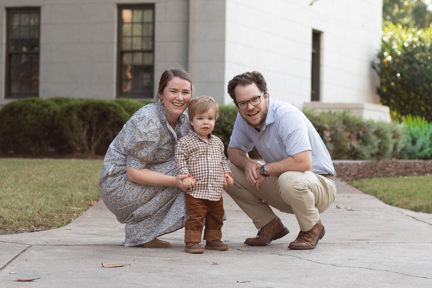 Family of three posing together on sidewalk in front of white building.
