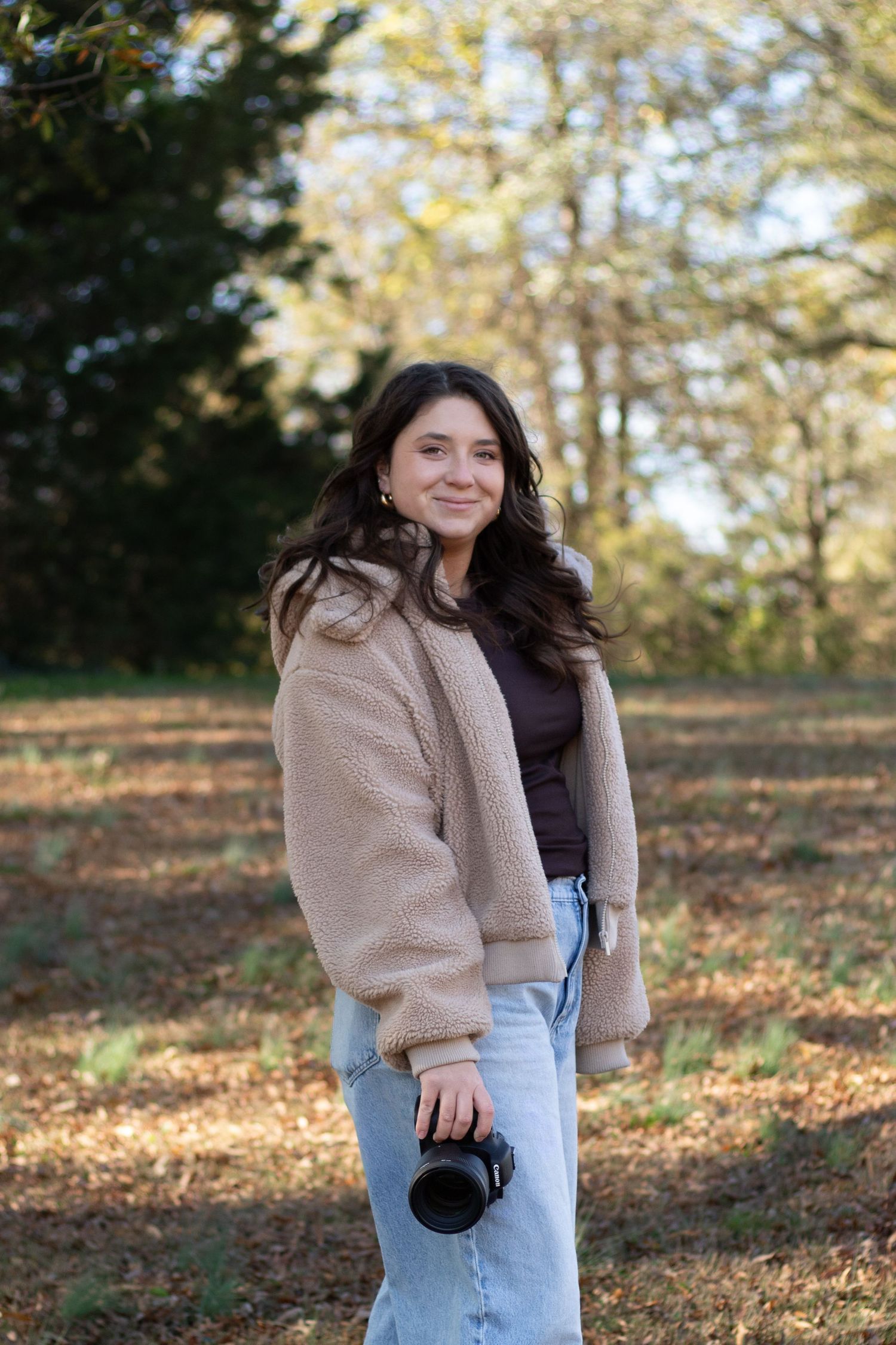 Woman in beige coat and jeans smiling while holding camera in autumn forest.