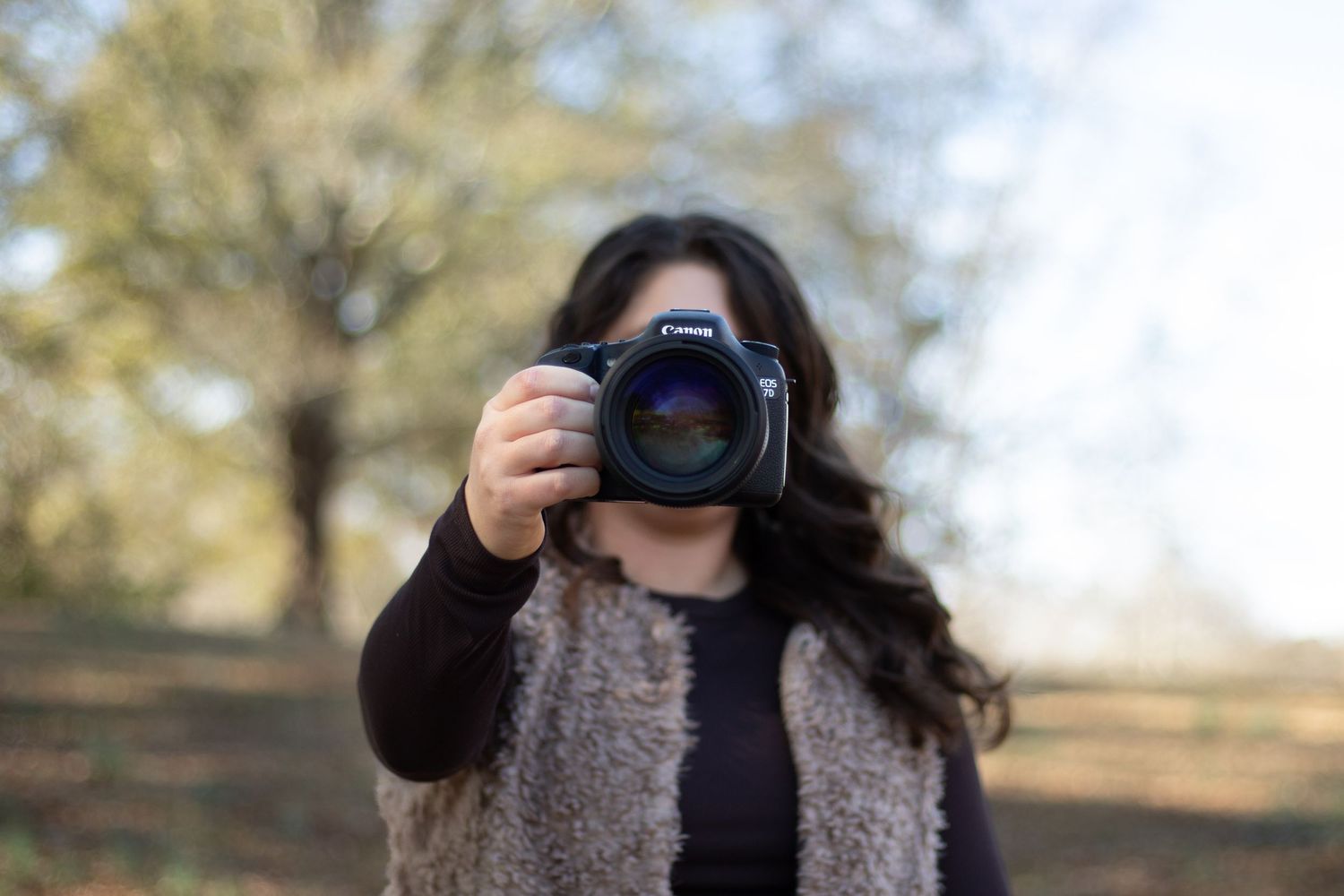Person holding camera up to face in outdoor autumn setting with trees in background.