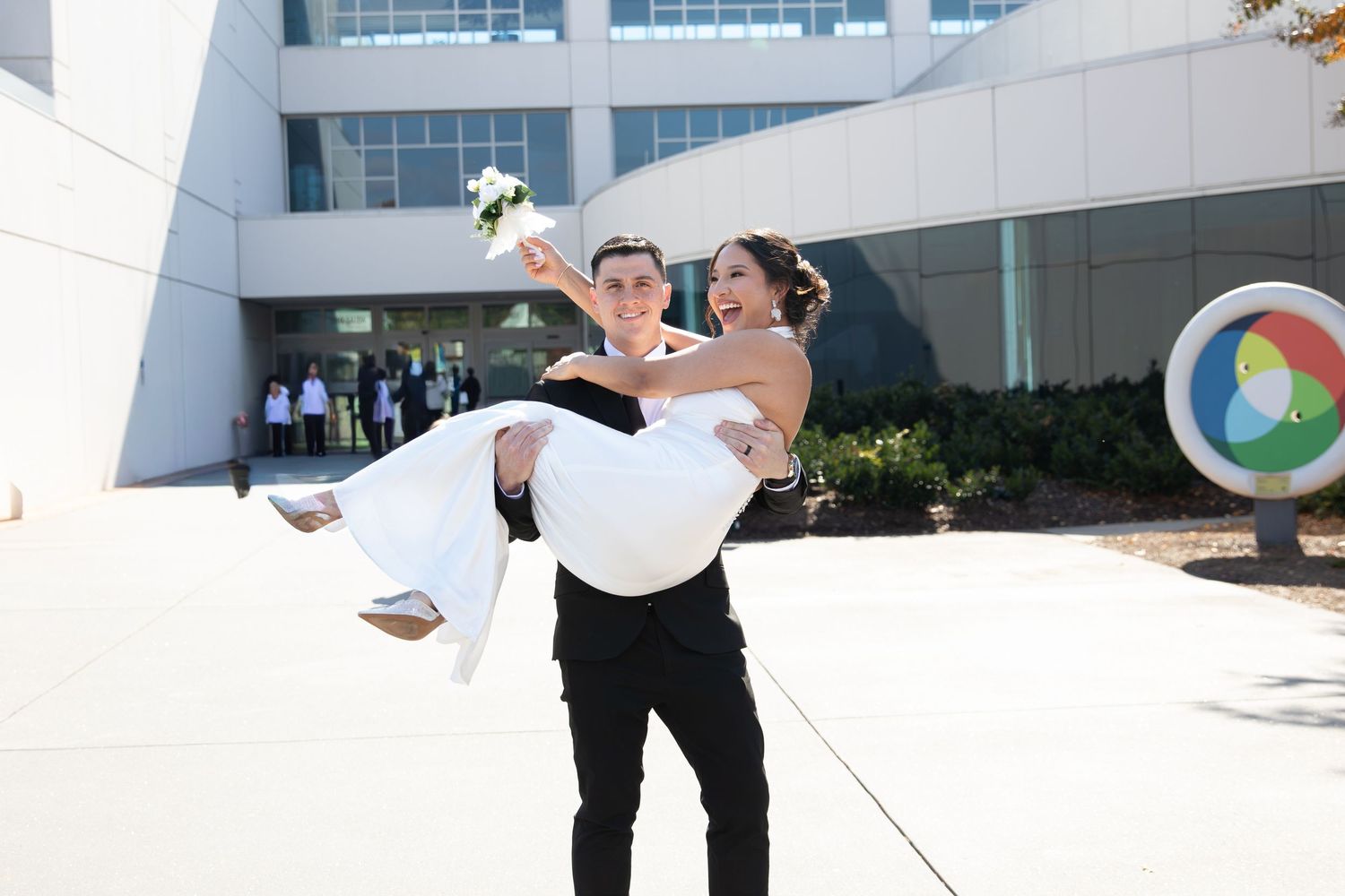 A couple poses together in formal wedding attire in front of a modern building.
