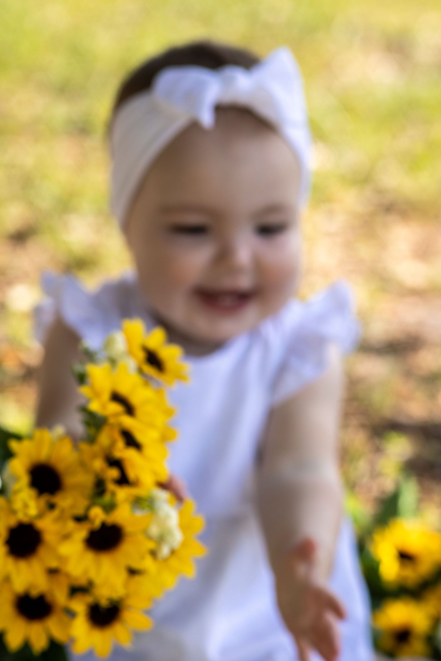 Smiling baby girl in white dress and bow headband holding yellow sunflowers outdoors in a sunny garden.