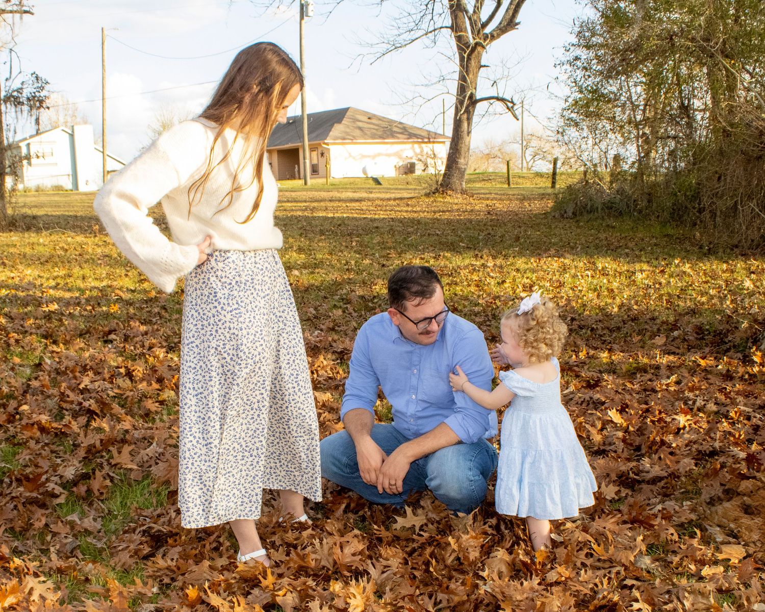 Family of three enjoying autumn leaves outdoors, with parents and toddler girl playing in a yard full of fallen leaves.