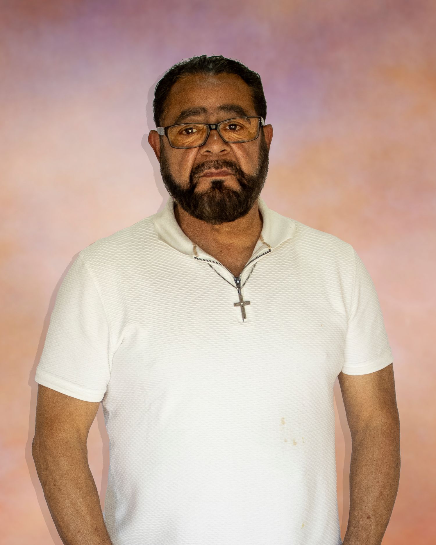 Middle-aged man with beard and glasses wearing white polo shirt and cross necklace poses against colorful studio backdrop.