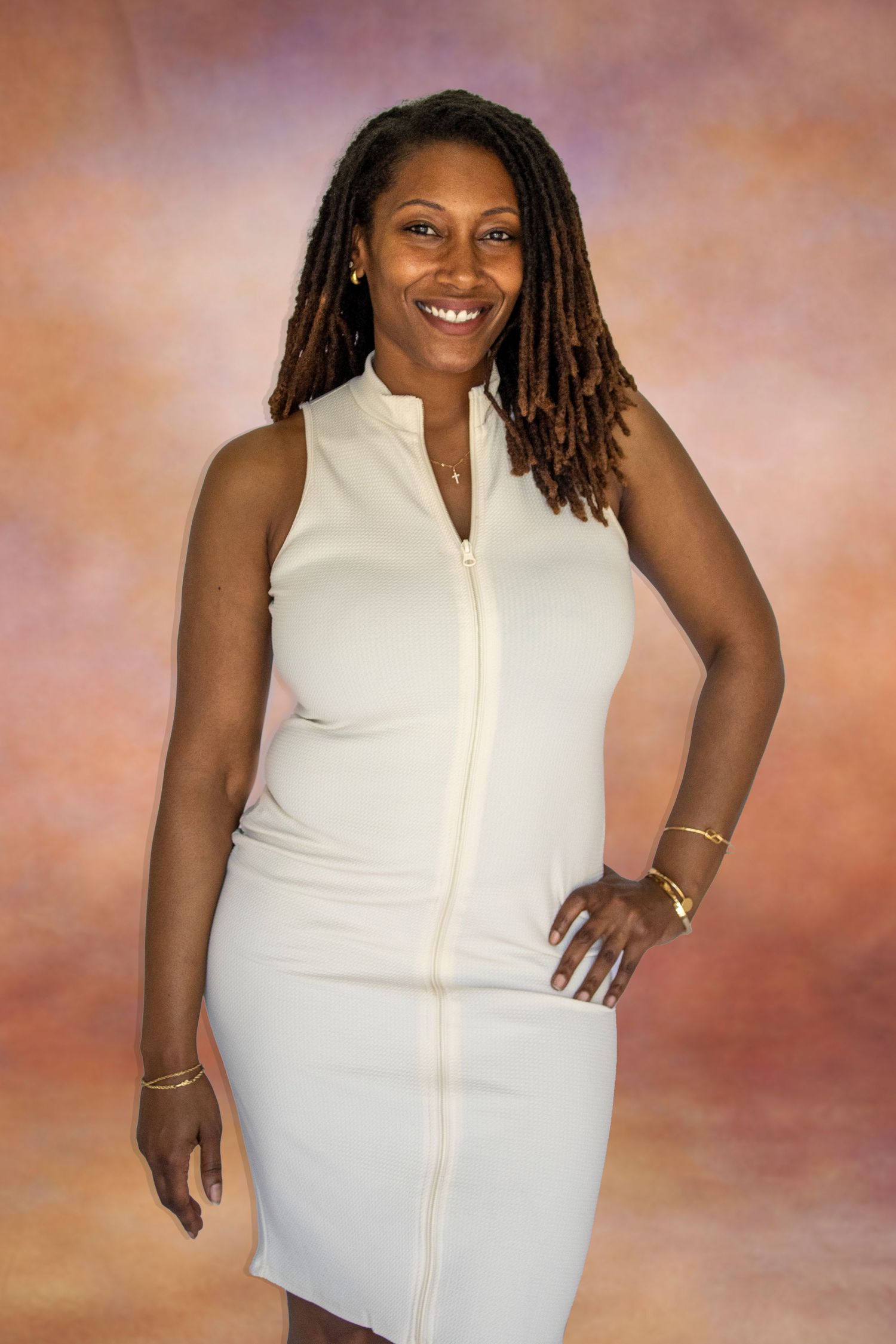 Smiling woman with locs wearing a white sleeveless zip-front dress and gold bracelets poses against a colorful backdrop.