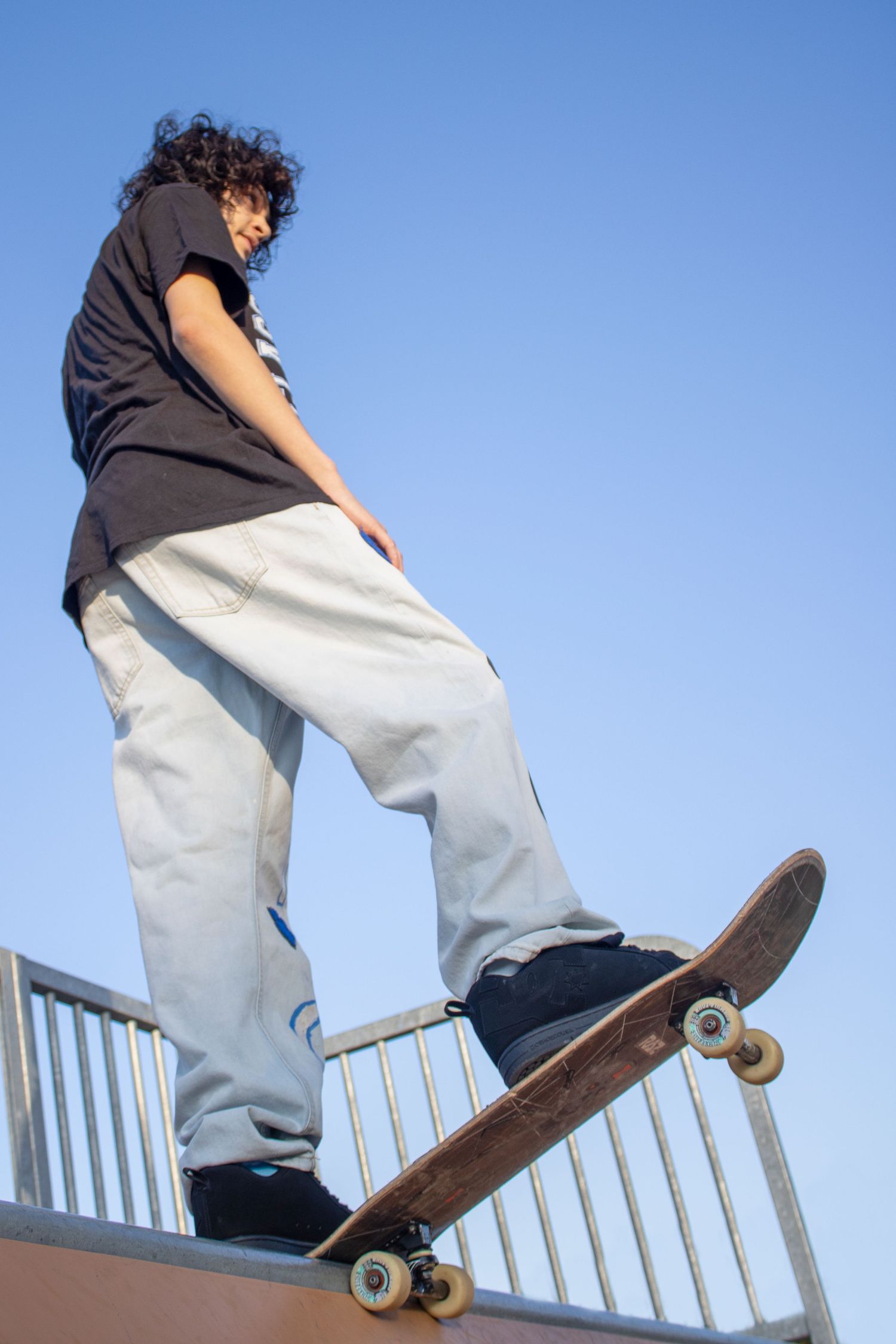 Young skateboarder in baggy jeans stands atop a skate ramp with skateboard, blue sky background.
