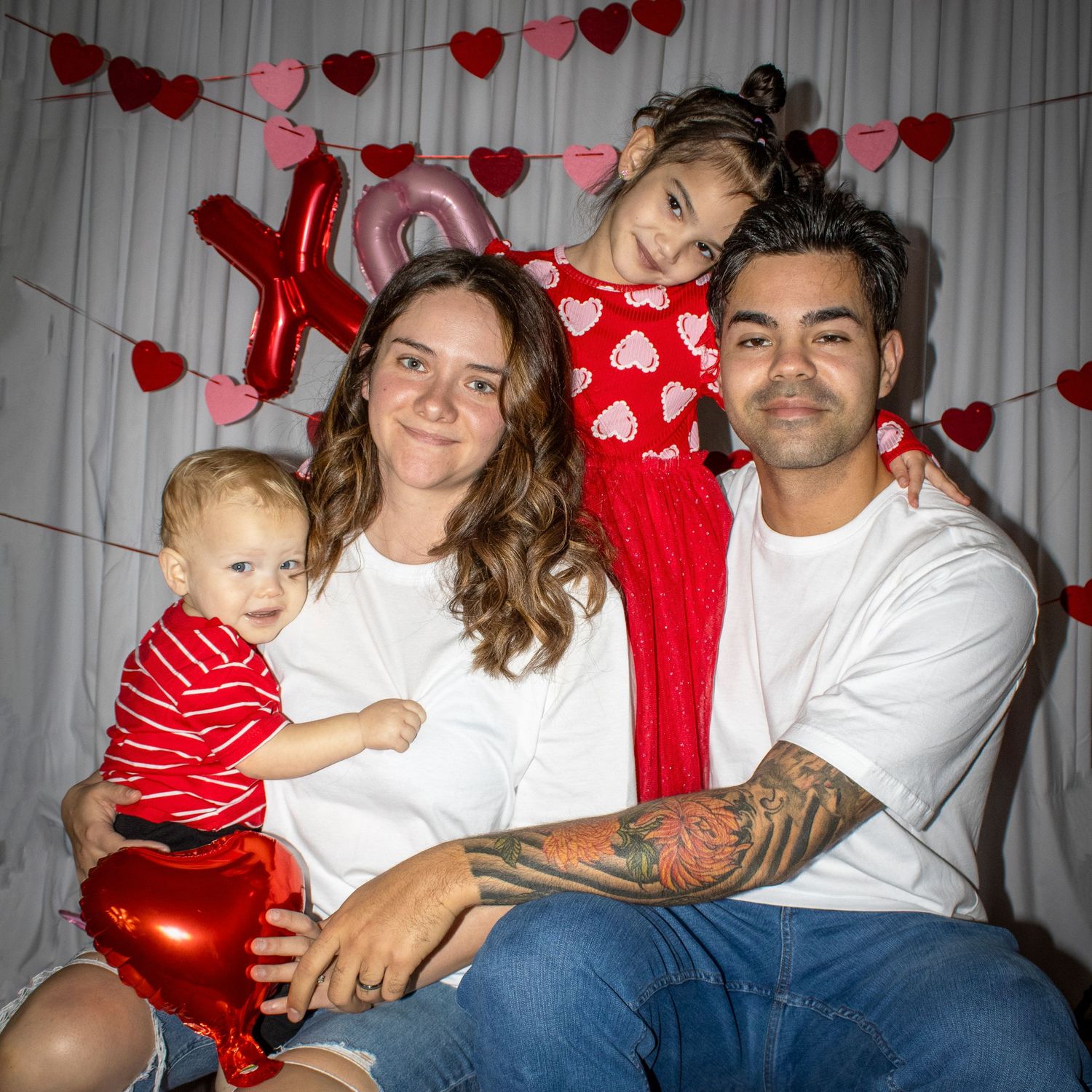 Family Valentine's Day photo with parents, toddler, and young girl posing with red heart balloons and XO decorations.