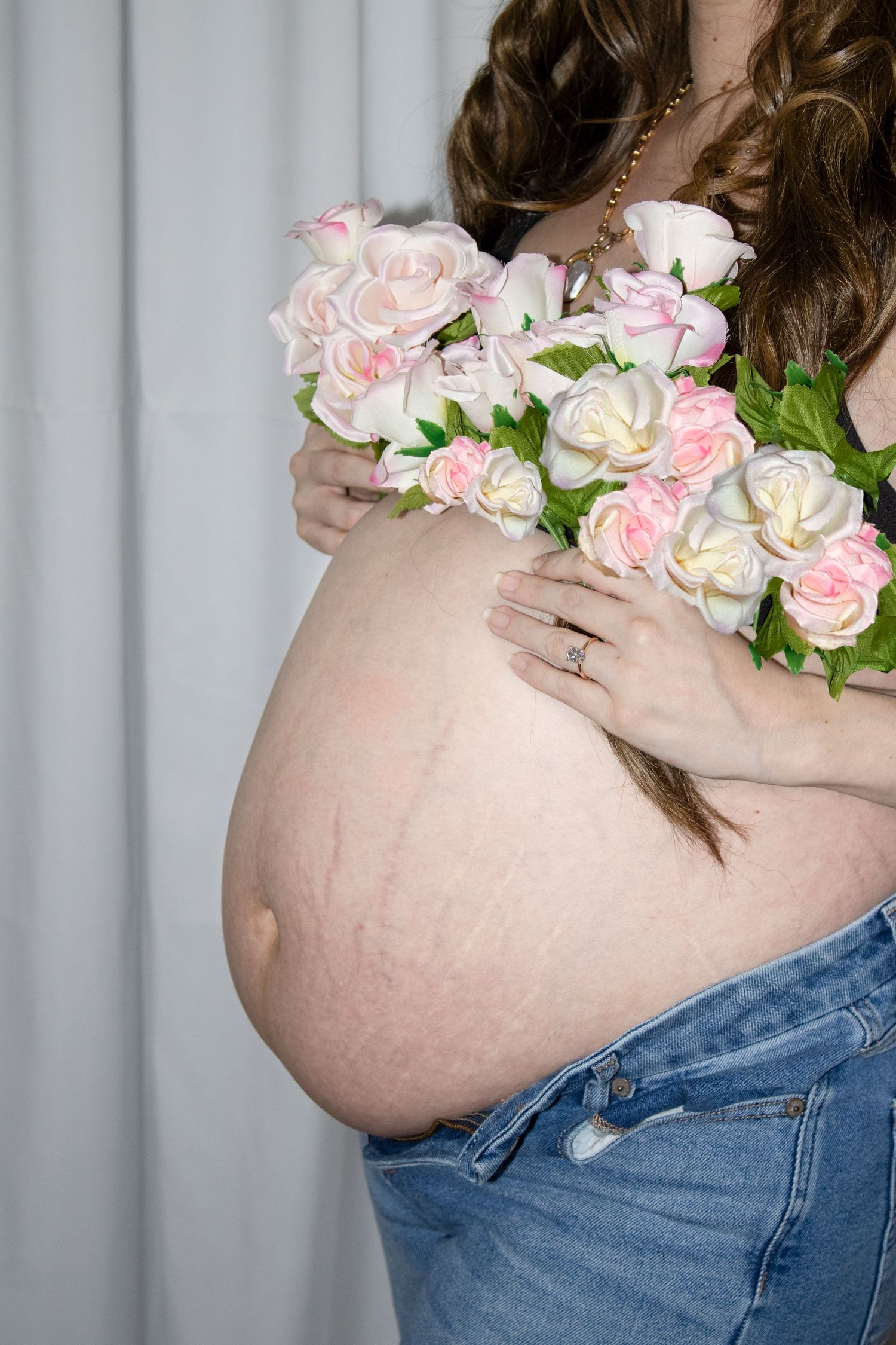 Pregnant woman holding pink and white roses bouquet, showing bare baby bump with stretch marks, wearing blue jeans.