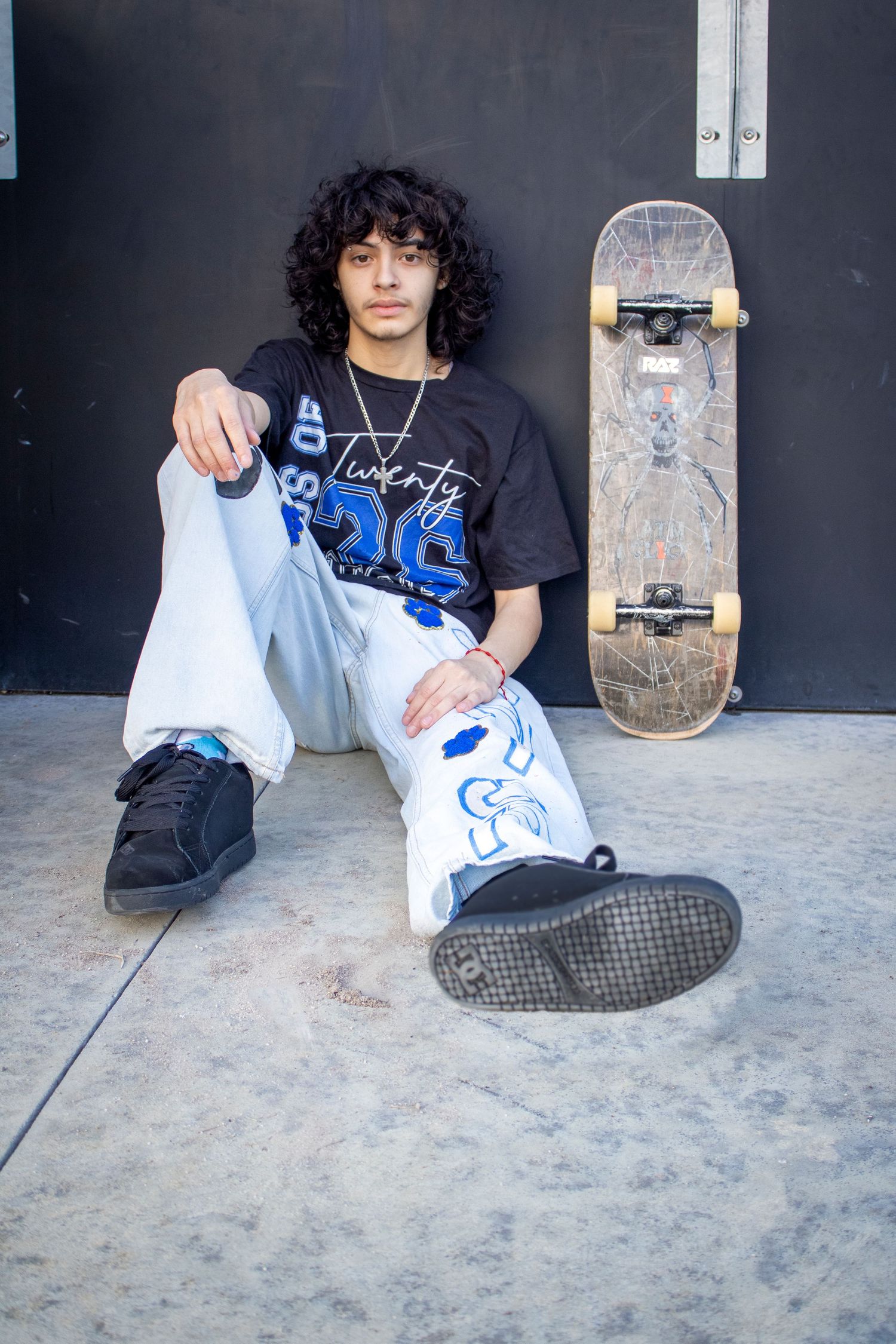 Young skater with curly hair sits against dark wall in graphic tee and white pants beside a worn skateboard.