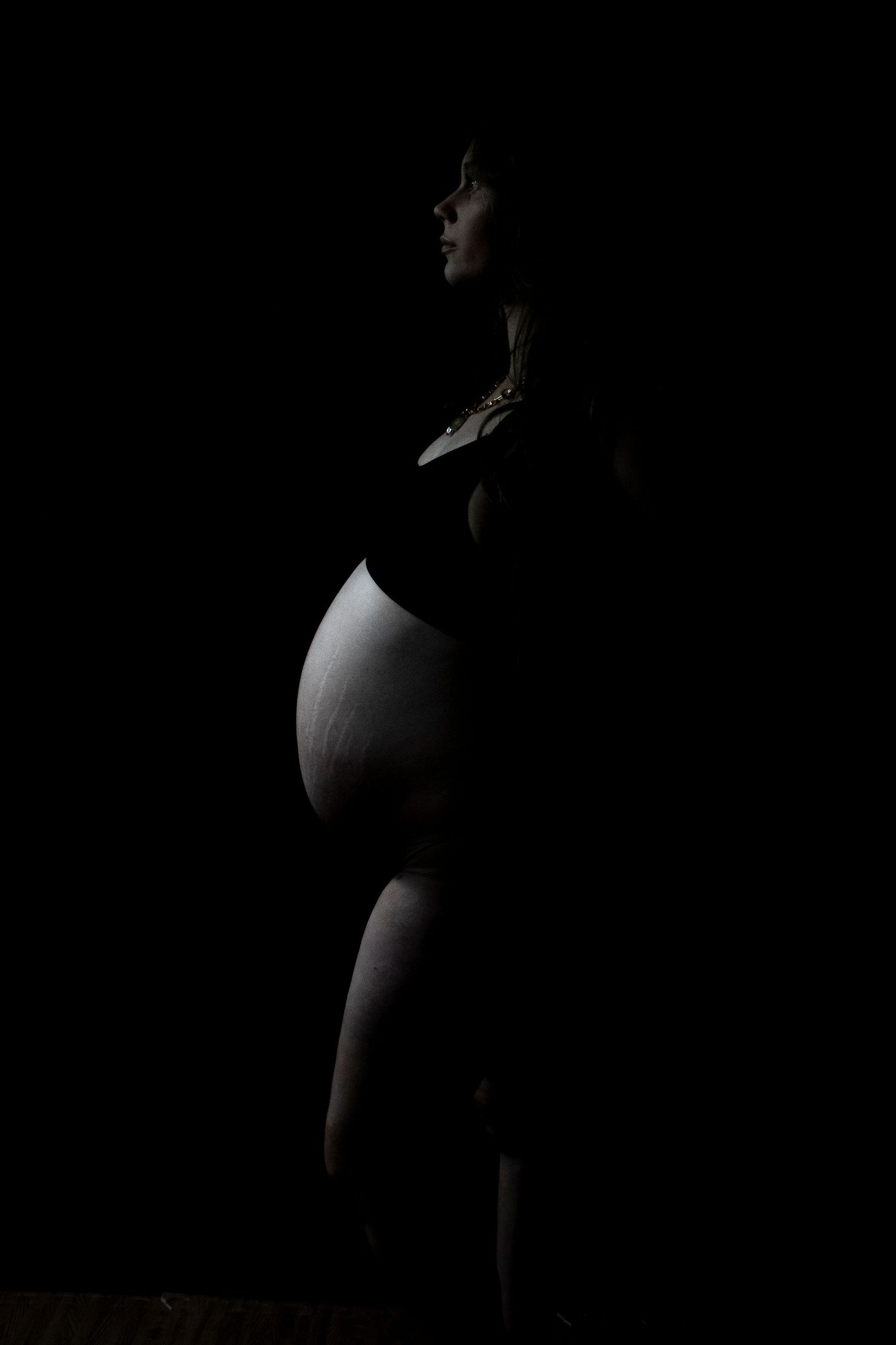 Silhouette of a pregnant woman standing against a dark background, highlighting her baby bump in dramatic low-key lighting.