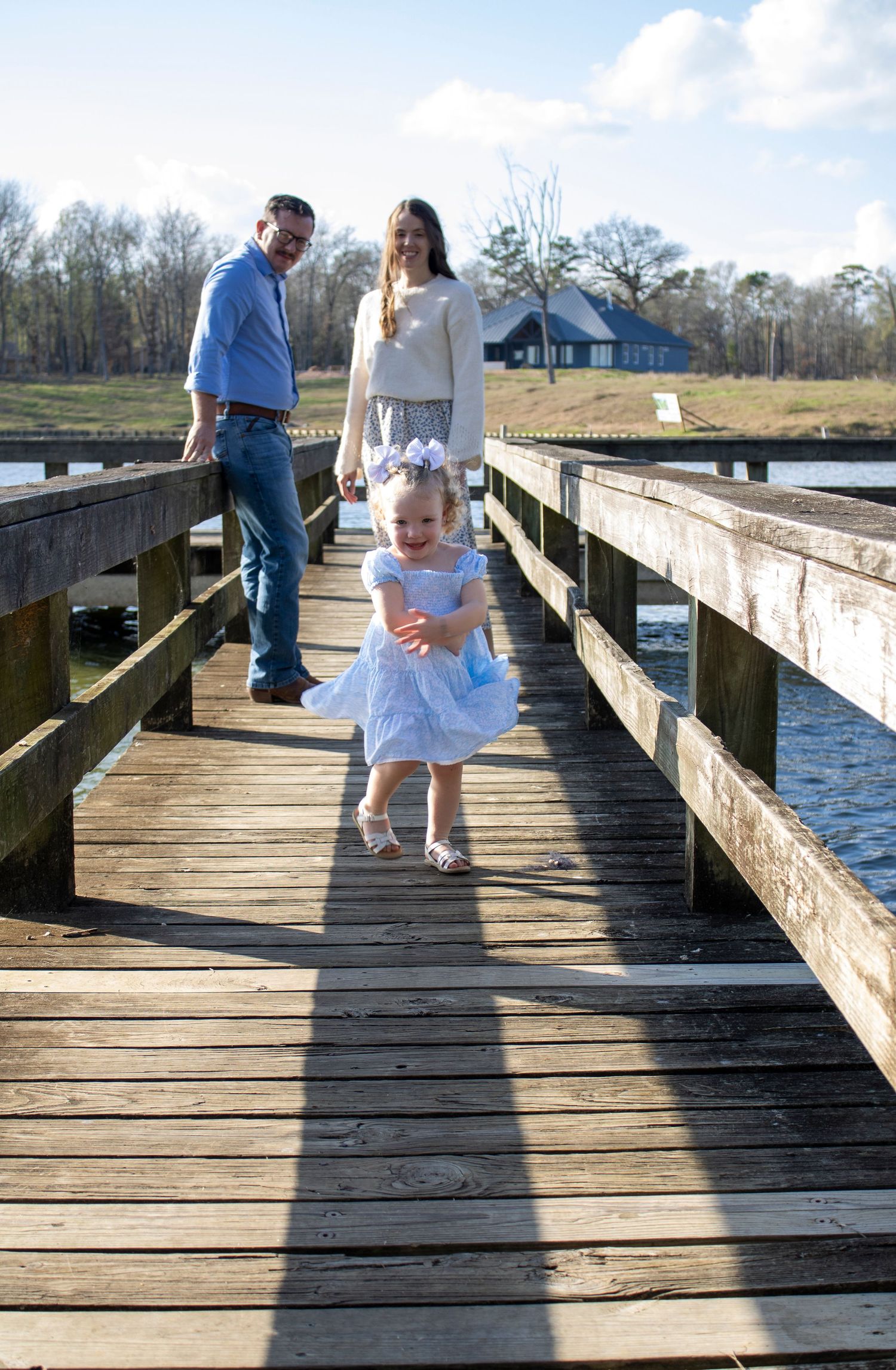 A young girl in a blue dress walks on a wooden dock while her parents watch, with a lake and house in the background.