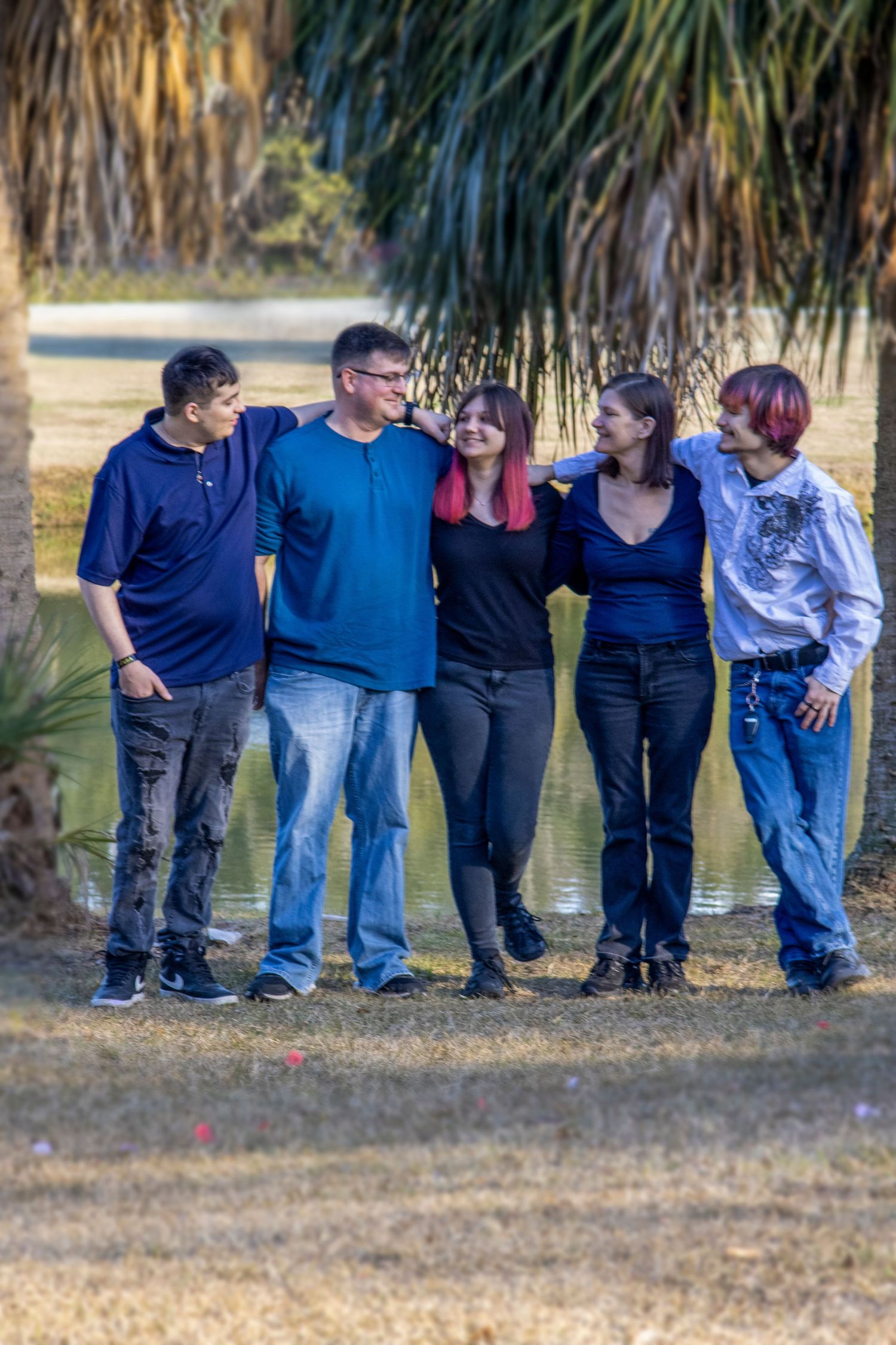 Five people smiling and embracing outdoors near a pond surrounded by palm trees on a sunny day.