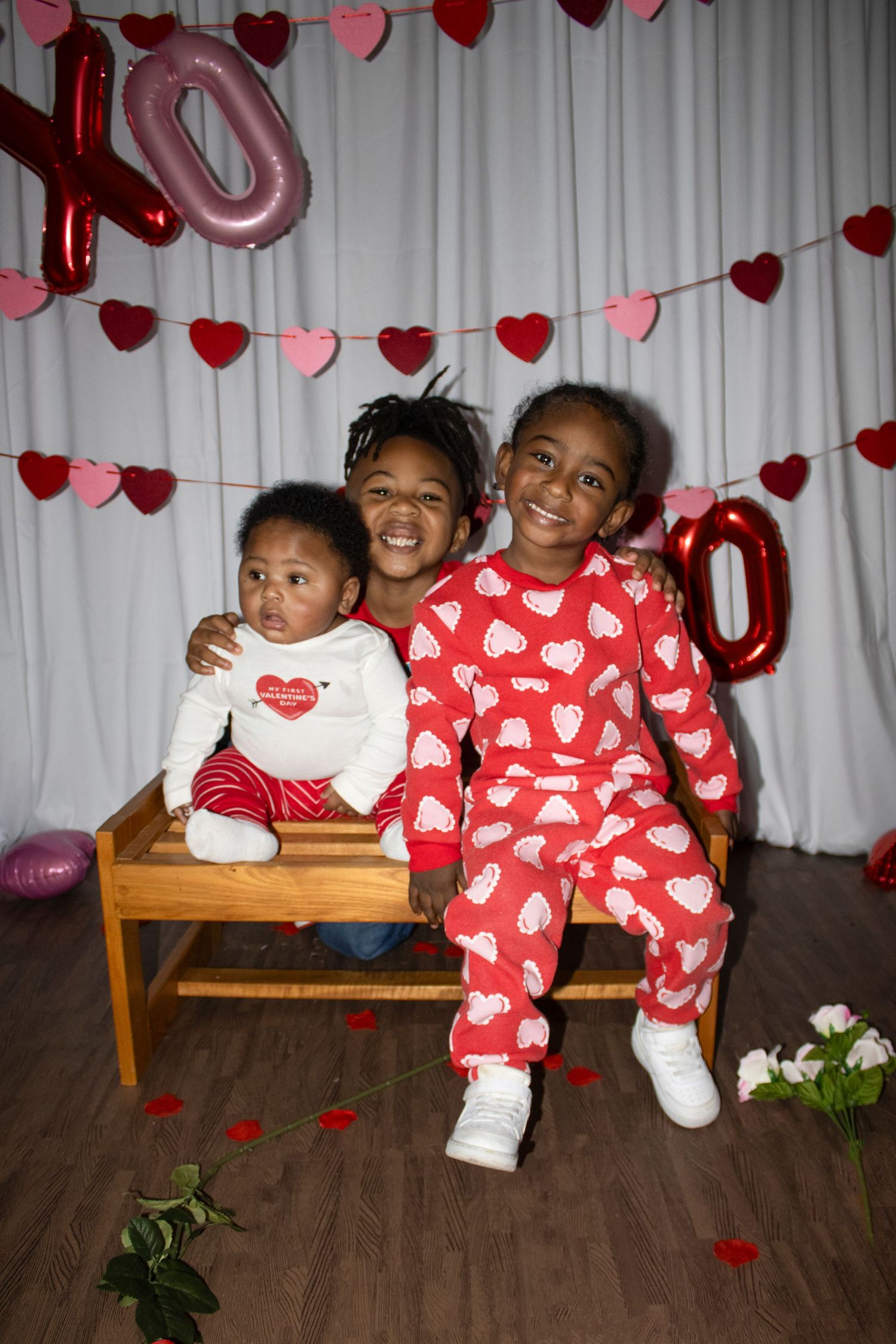 Three children pose on a bench with Valentine's Day heart decorations and XO balloons in the background.