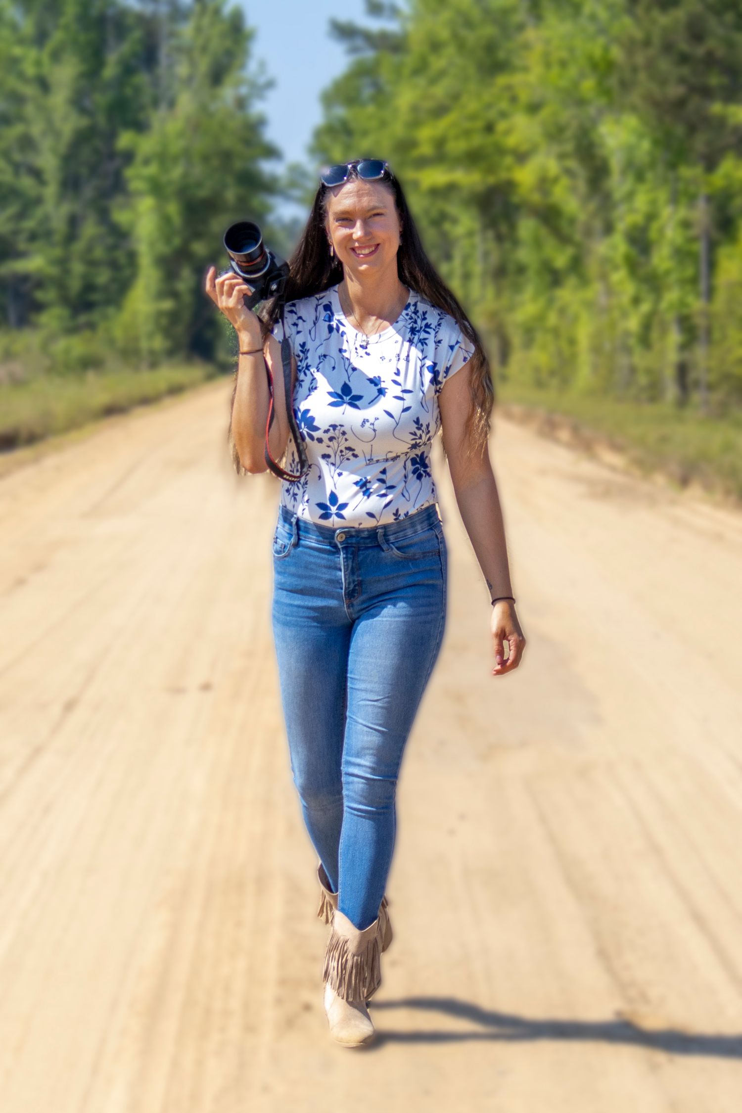 Woman in blue jeans and floral top walks down a sunny dirt road holding a camera with trees in background.