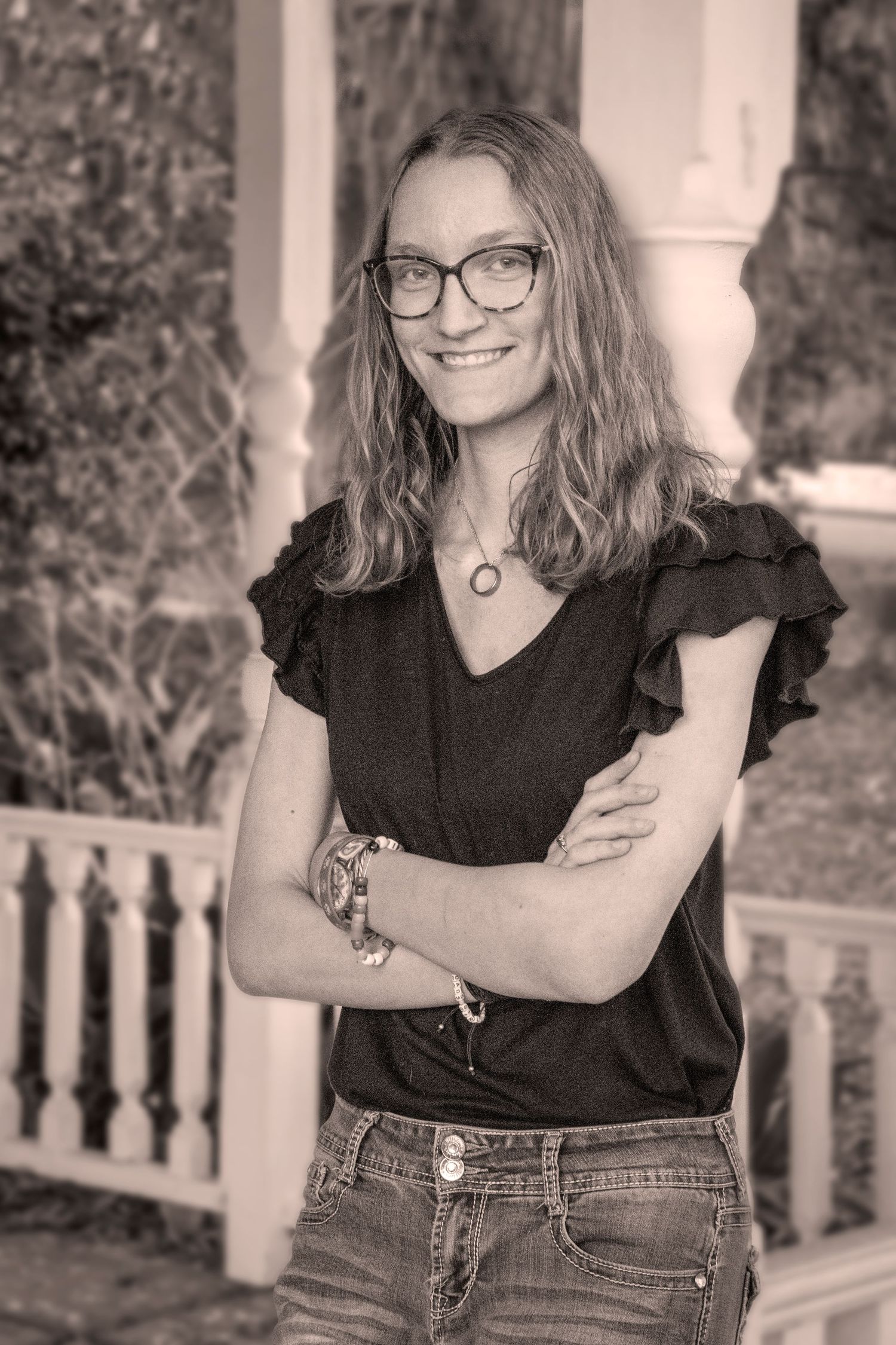 Person in black top and glasses poses confidently against white porch railing.