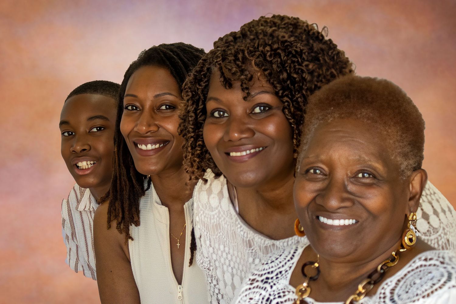 Four generations of Black women and a young boy smile together in a warm studio family portrait.