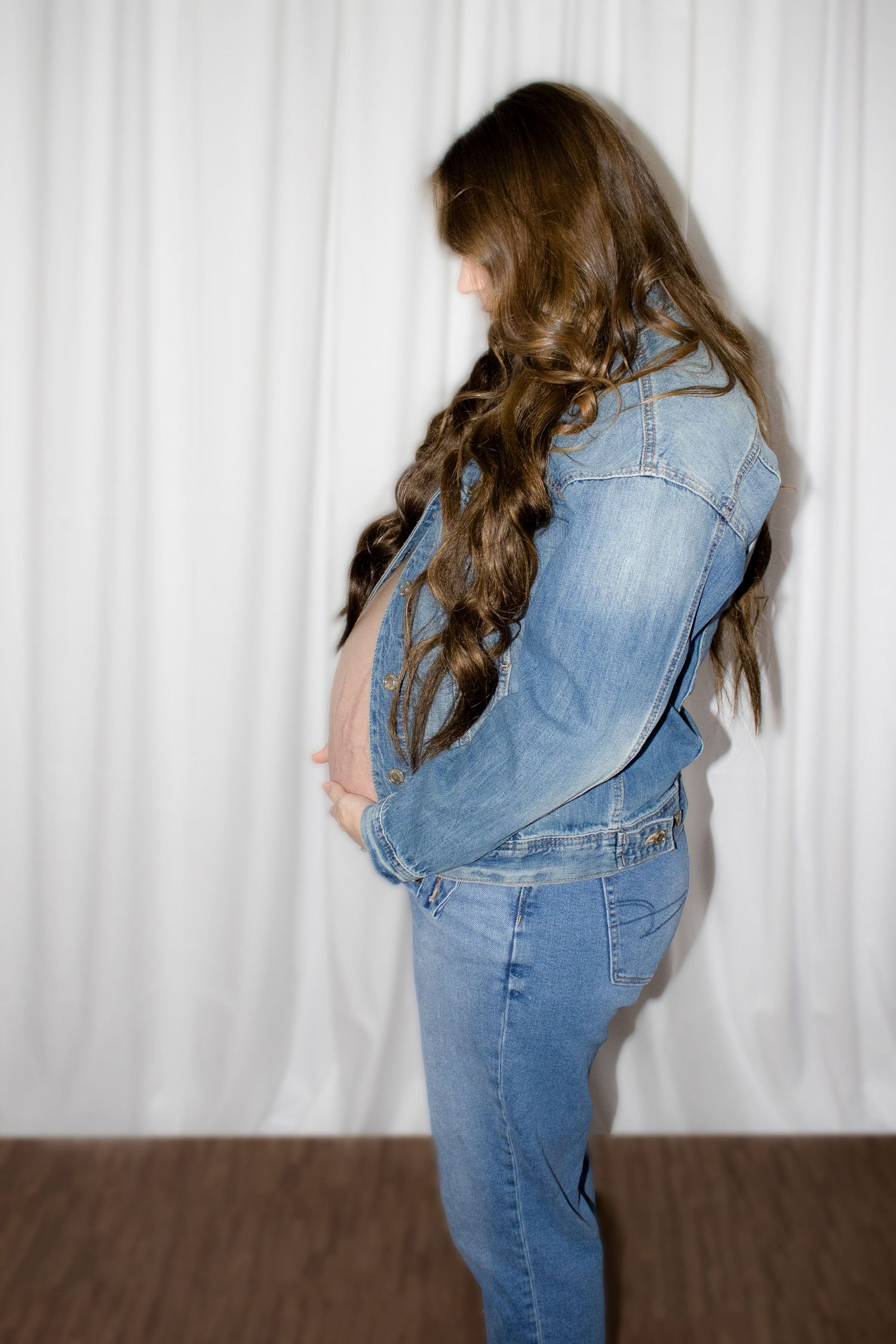 Pregnant woman in denim jacket and jeans holding baby bump, long wavy brown hair, standing against white curtain.