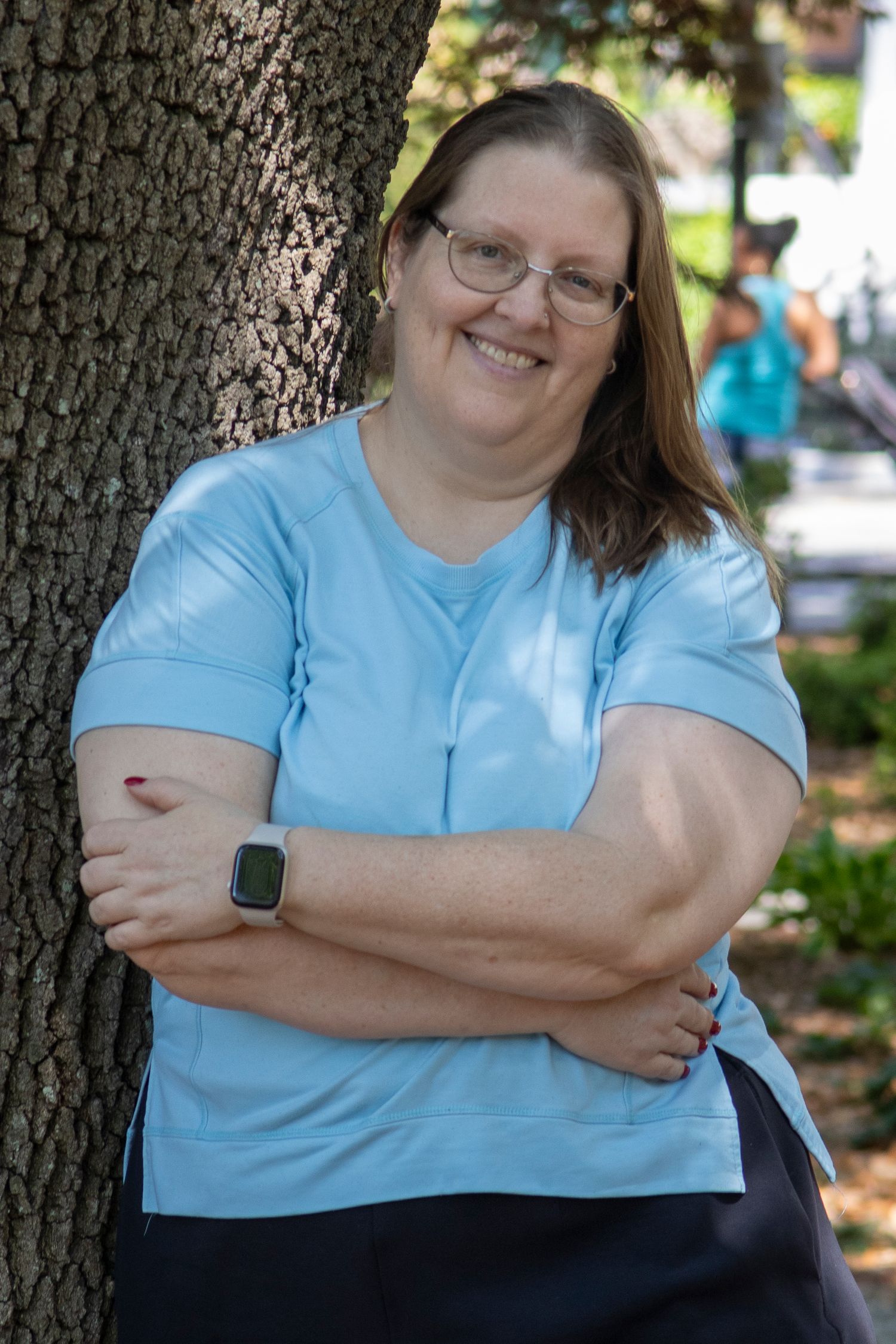 Smiling woman in light blue top leaning against tree with arms crossed, wearing glasses and a smartwatch outdoors.