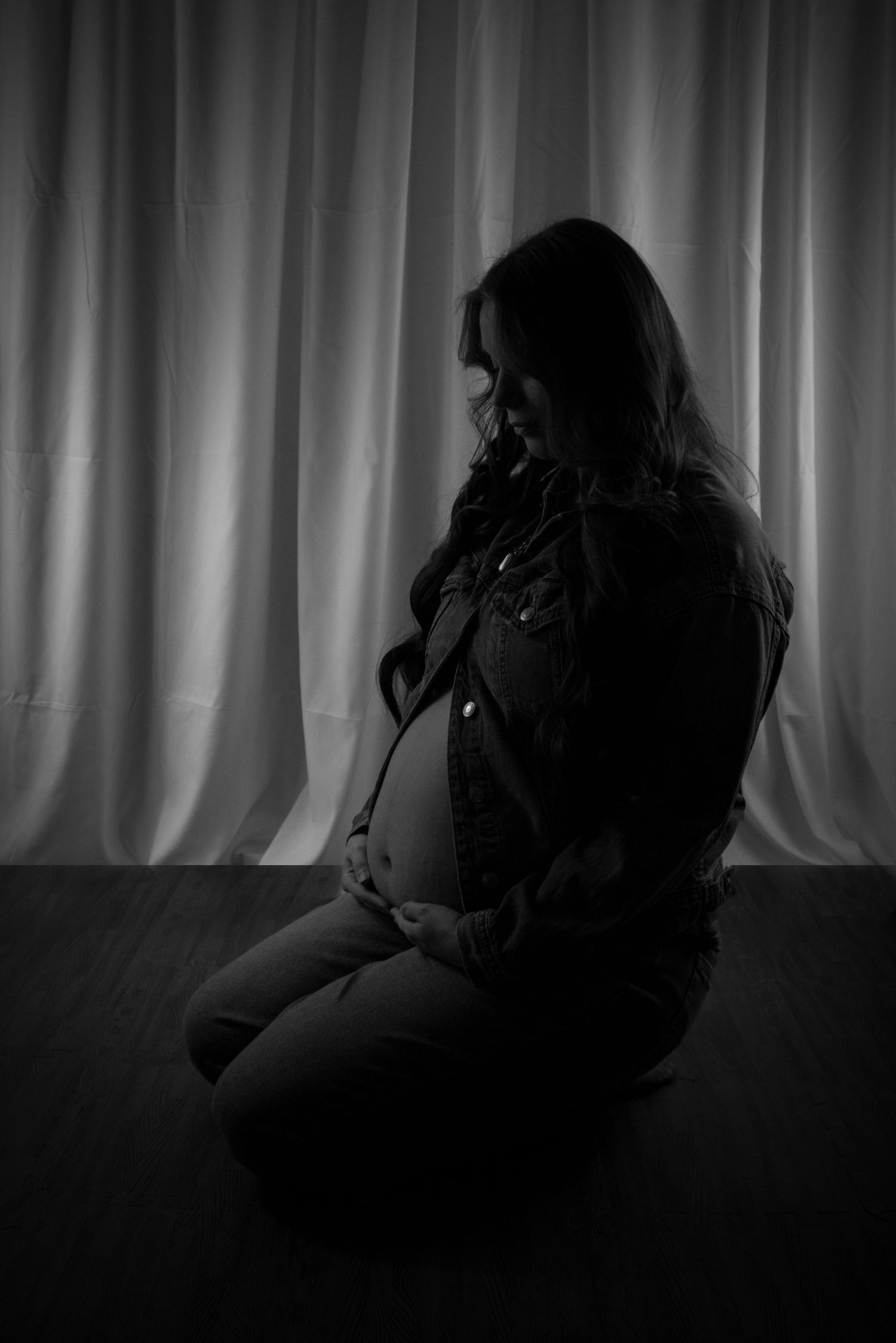Black and white silhouette of a pregnant woman sitting on floor cradling her baby bump against draped curtains.