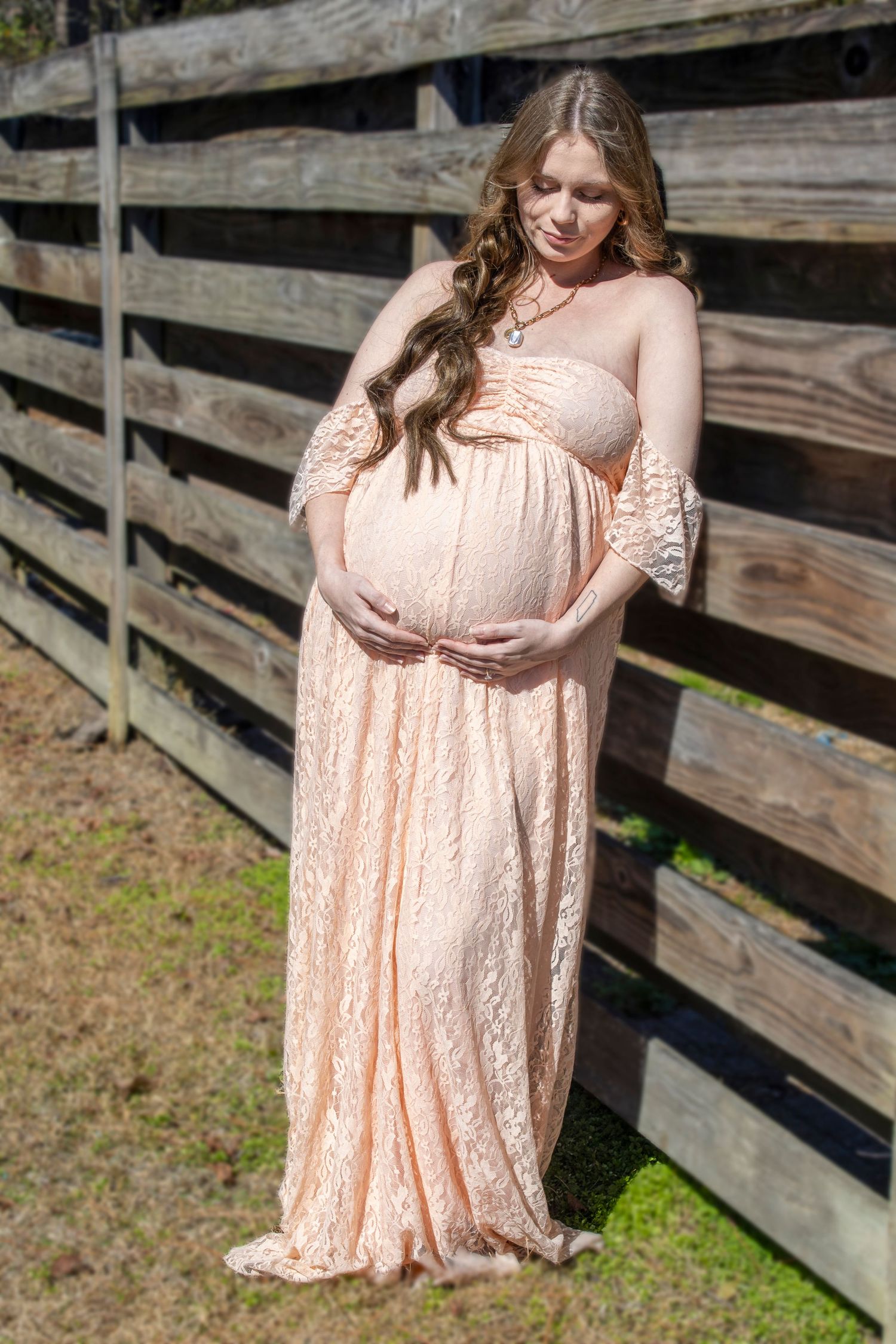 Pregnant woman in peach lace maternity gown cradling her baby bump outdoors near a rustic wooden fence.