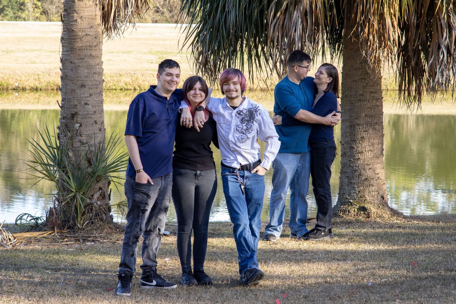 Family of five posing outdoors near a pond with palm trees on a sunny day.