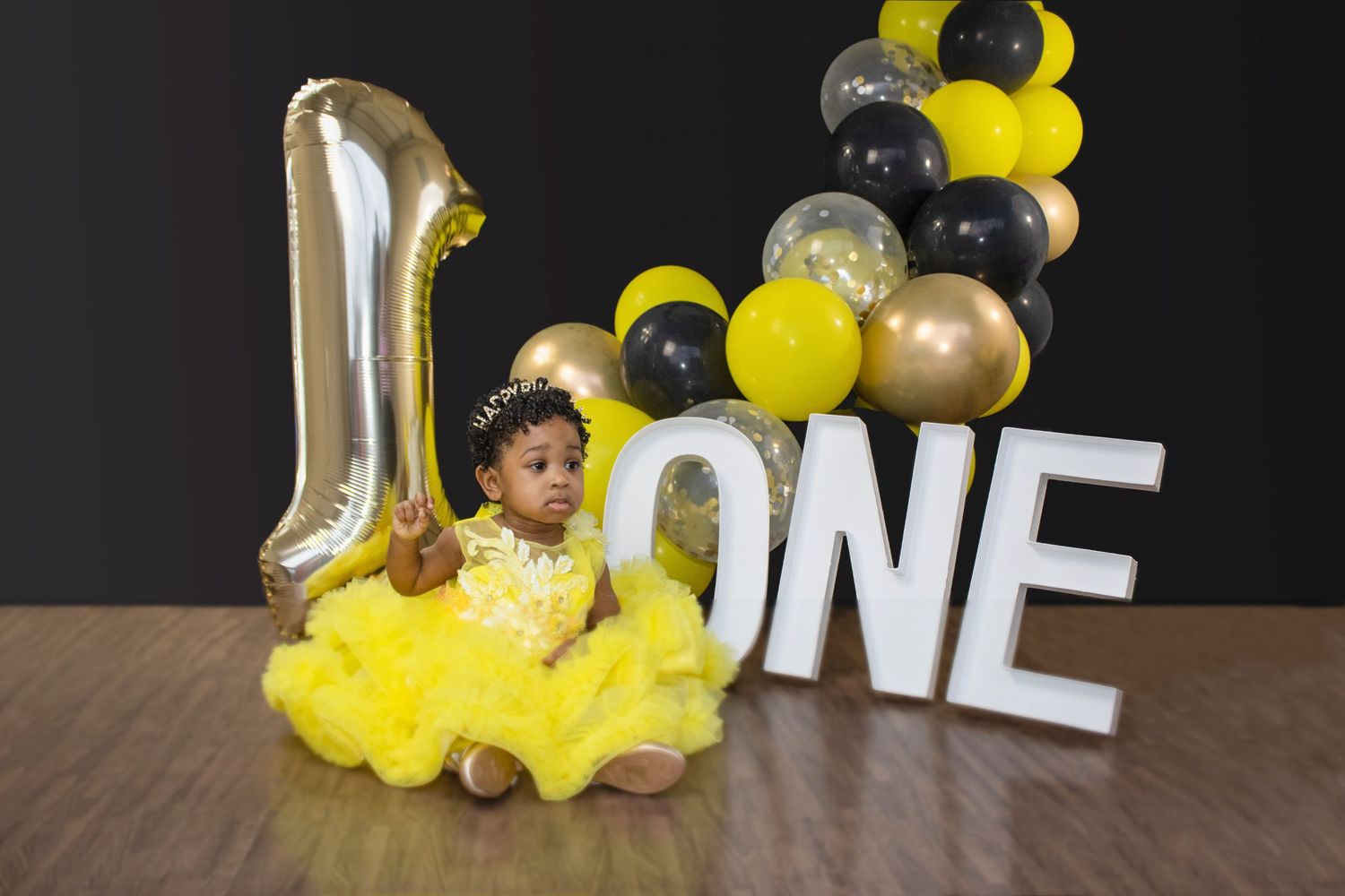 Baby girl in yellow tutu dress sits beside ONE sign with gold balloon number 1 and yellow black gold balloons for first birthday.