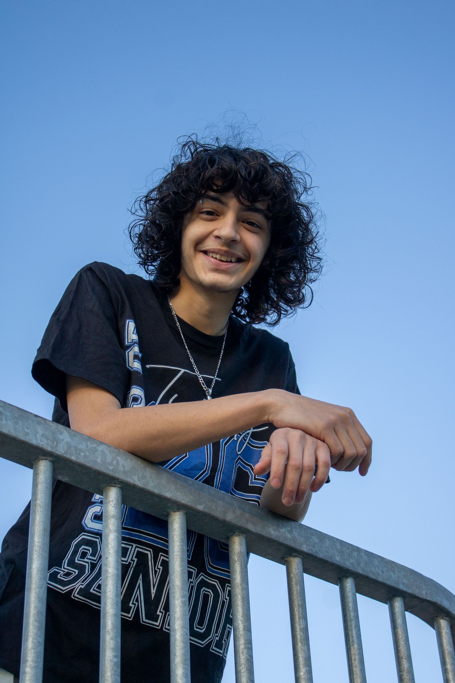 Smiling teen with curly black hair leans on metal railing wearing a black graphic tee against a blue sky.