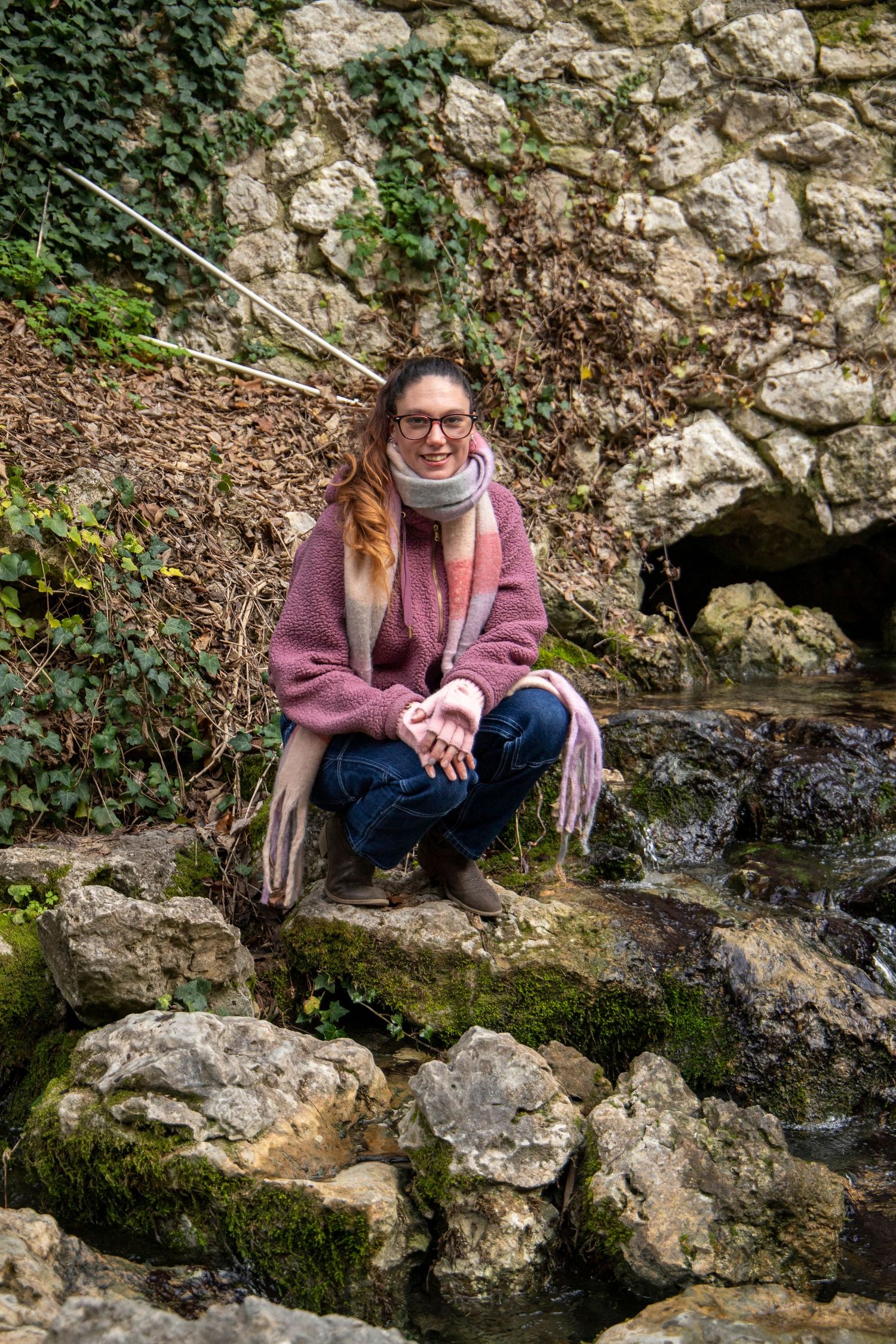 Person in a purple coat sitting on mossy rocks by a stream.