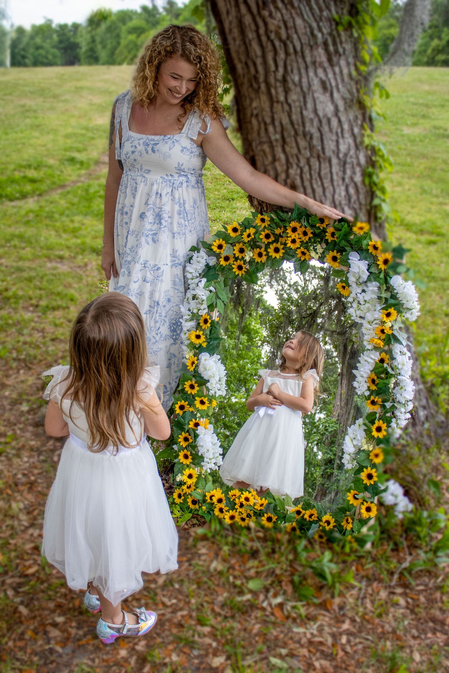 A mother holds a sunflower-framed mirror outdoors as her young daughter in white dress admires her reflection.