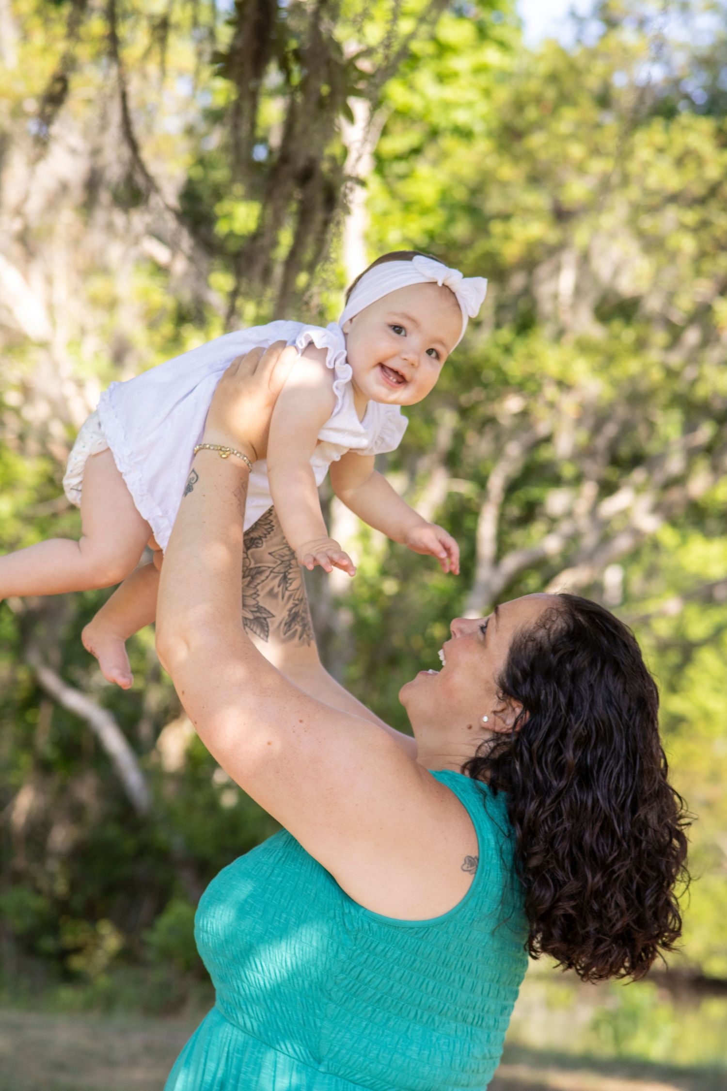 A joyful mother in a teal dress lifts her smiling baby girl in a white dress outdoors among lush green trees.