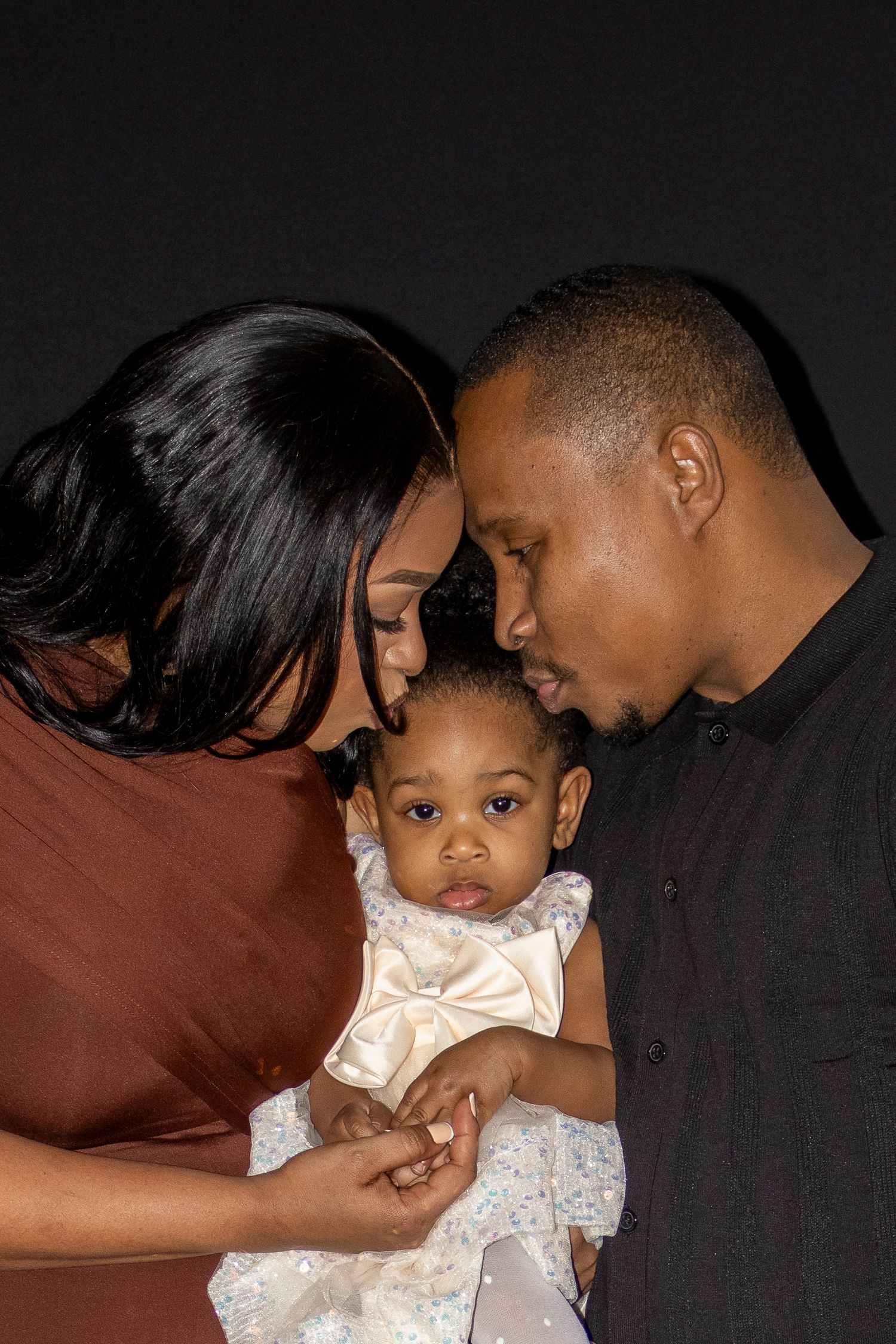 A loving family portrait of parents tenderly kissing their baby girl dressed in a white bow outfit against a dark background.