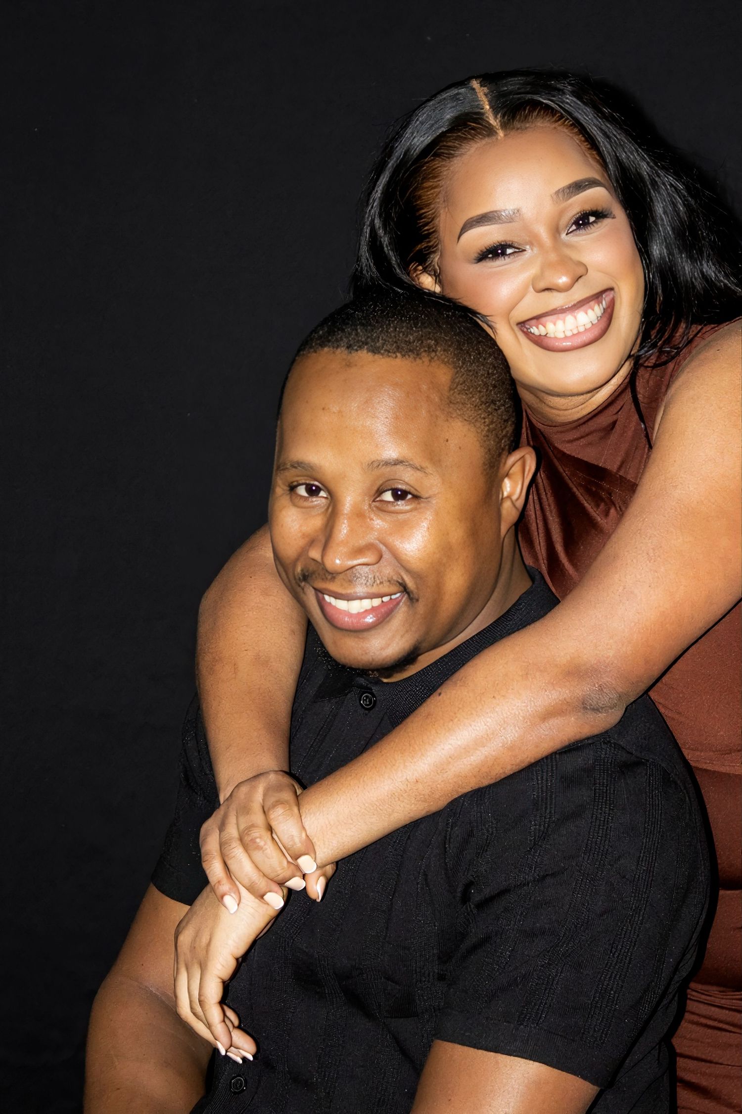 A joyful couple posing together against a dark background, woman embracing man from behind, both smiling warmly.