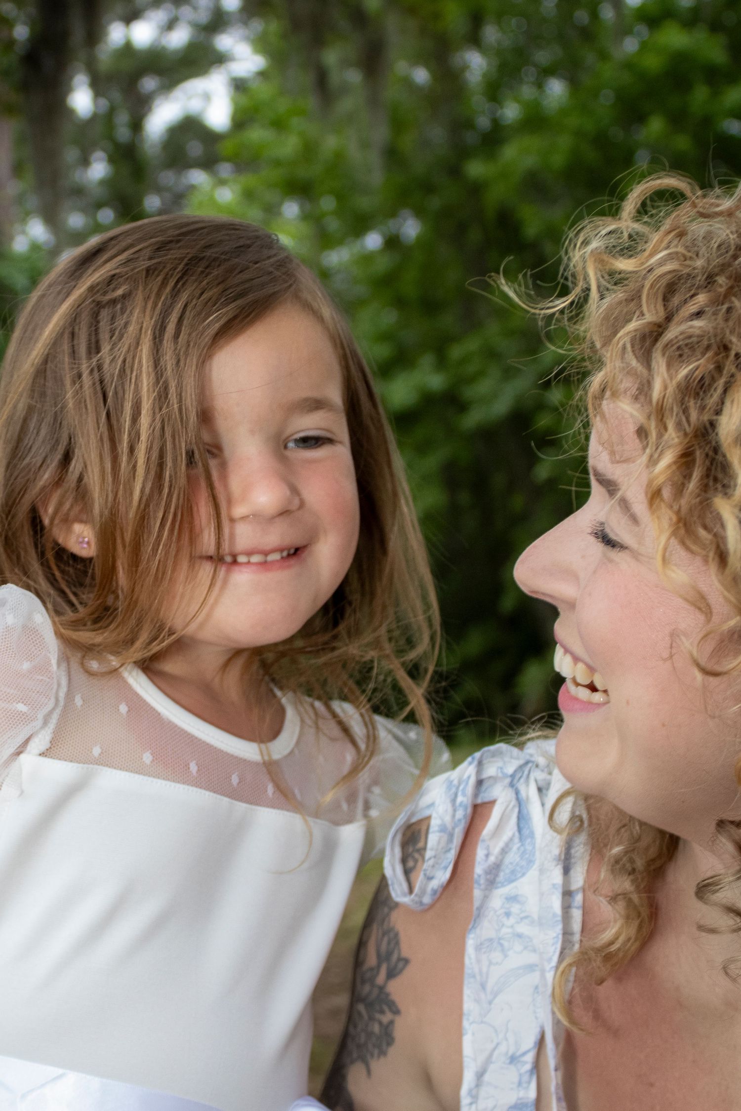 A young girl in a white dress smiles at a curly-haired woman outdoors, sharing a joyful mother-daughter moment.