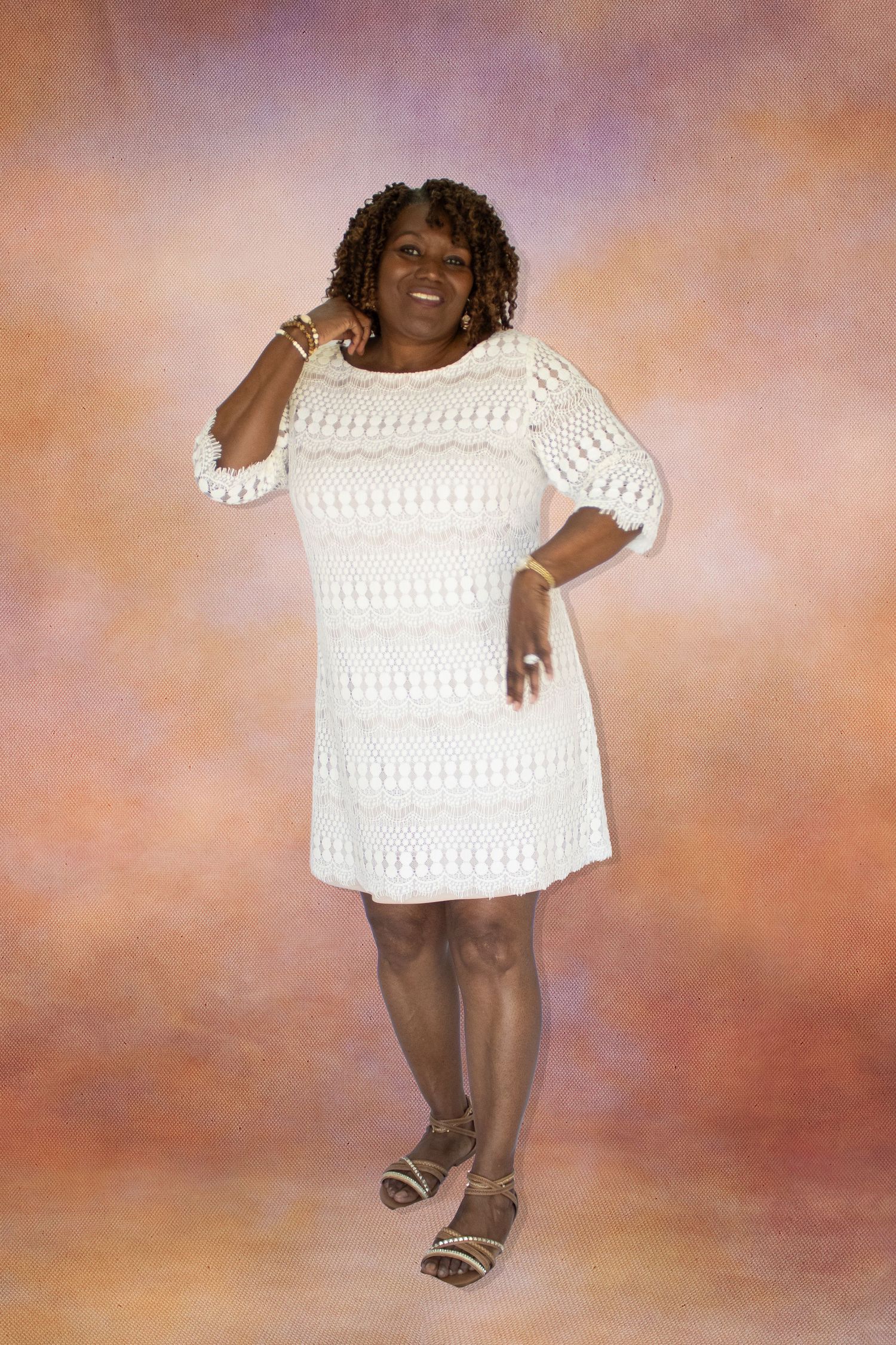 A smiling woman in a white crochet dress poses confidently against a soft pink and orange studio background.