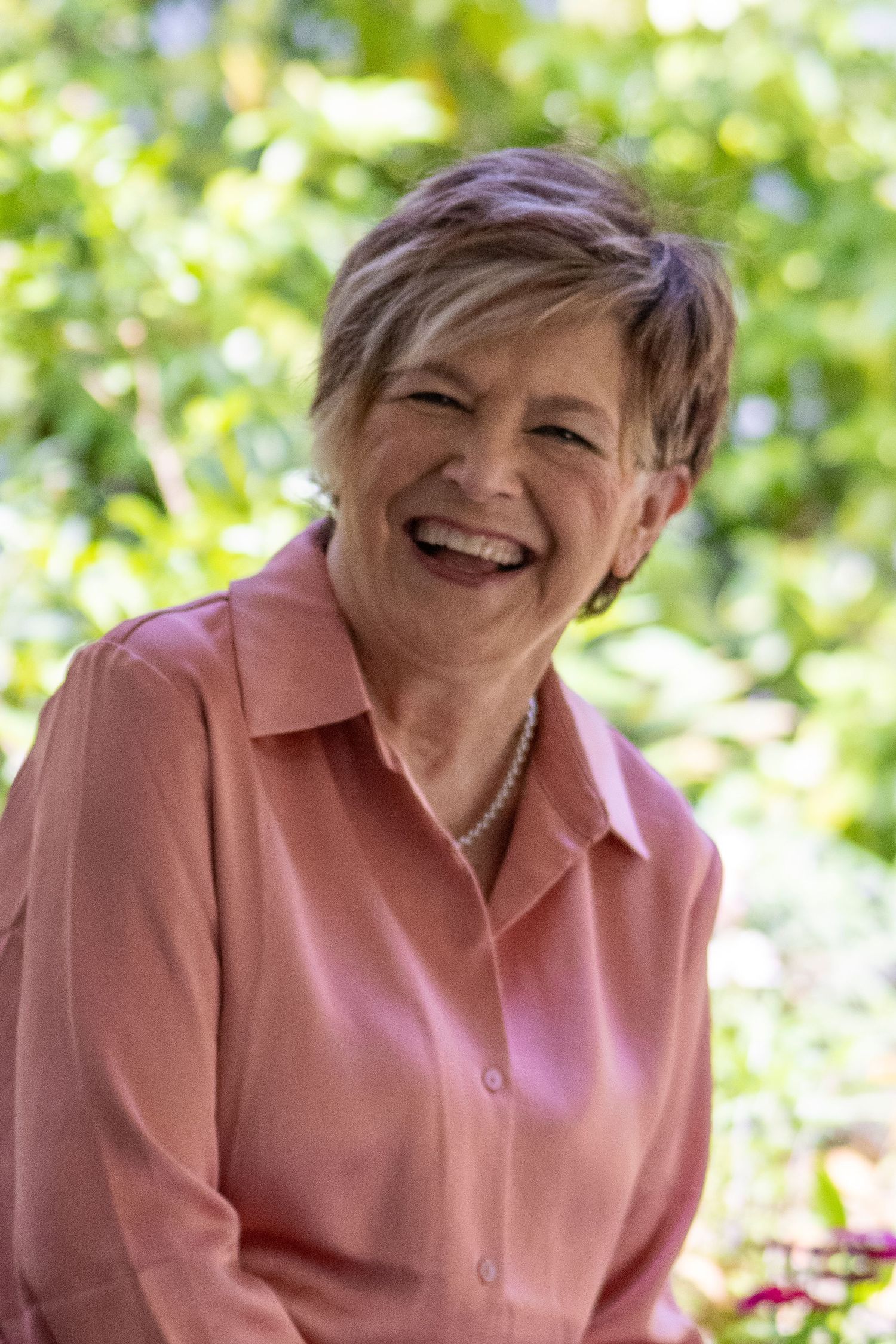 Smiling older woman with short hair wearing a pink blouse and pearl necklace, laughing outdoors in a lush green garden.