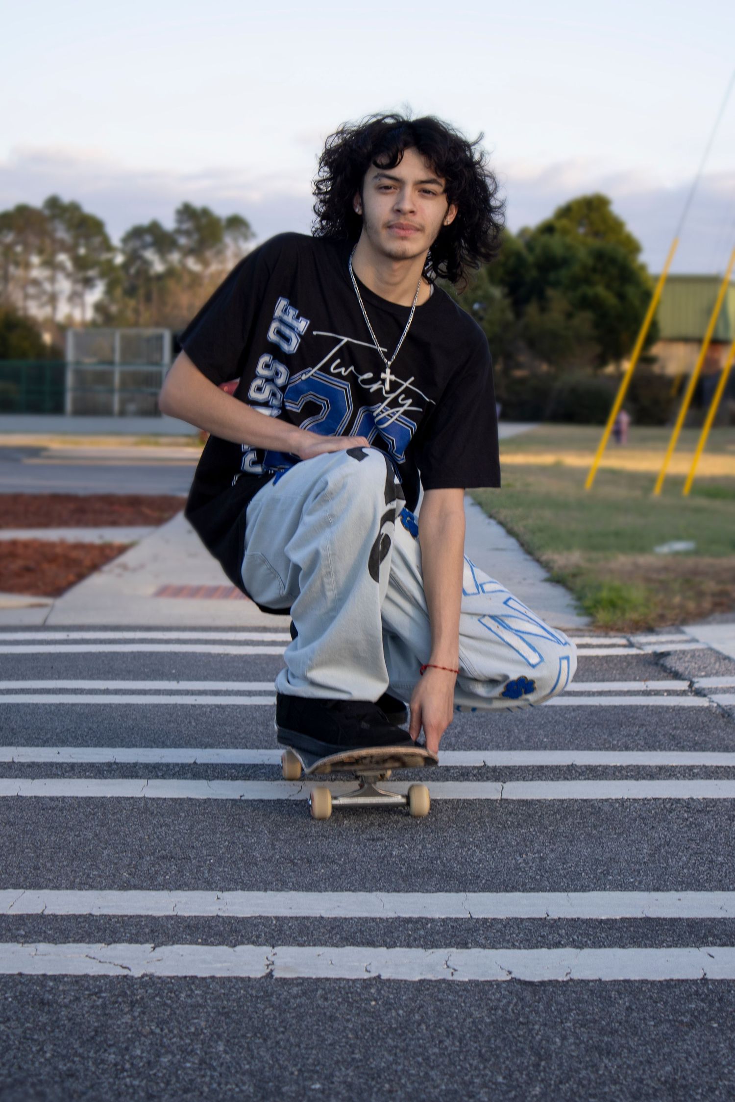 Young skateboarder with curly hair posing on skateboard at crosswalk in black graphic tee and baggy jeans.