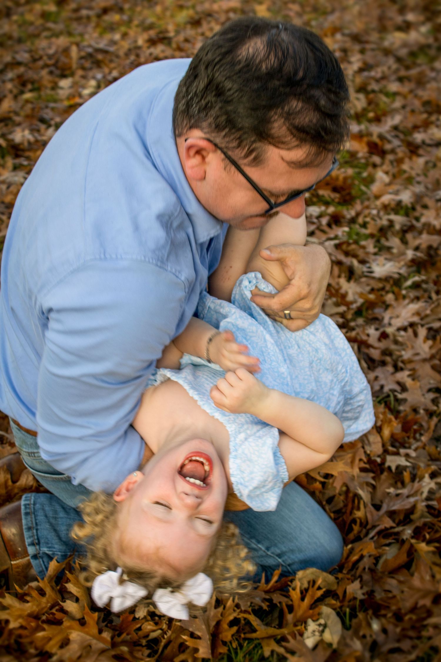 Father playfully tickling laughing toddler girl in autumn leaves during fun outdoor family photo session.
