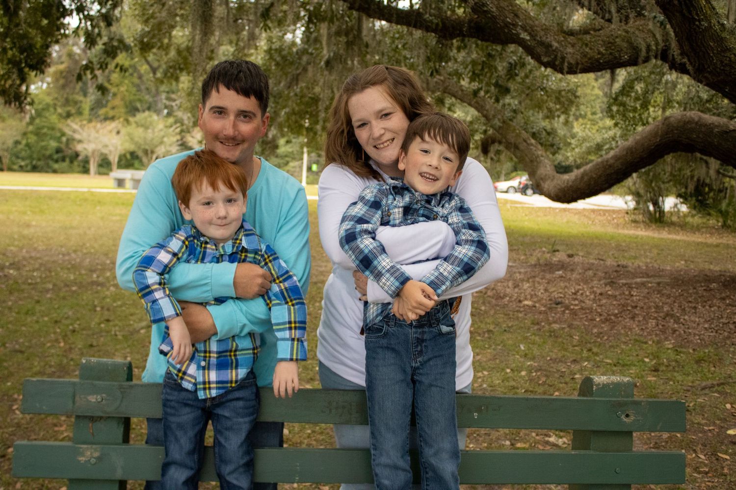 A family sits together on a wooden bench under a large oak tree on a sunny day.