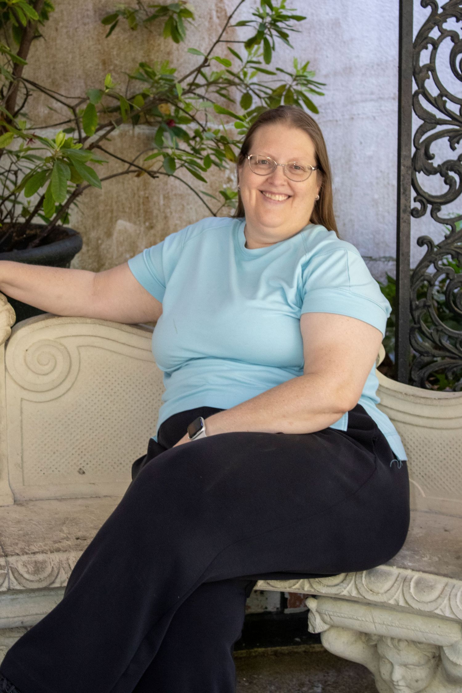 Smiling woman in light blue t-shirt and black pants sitting relaxed on ornate white bench outdoors.