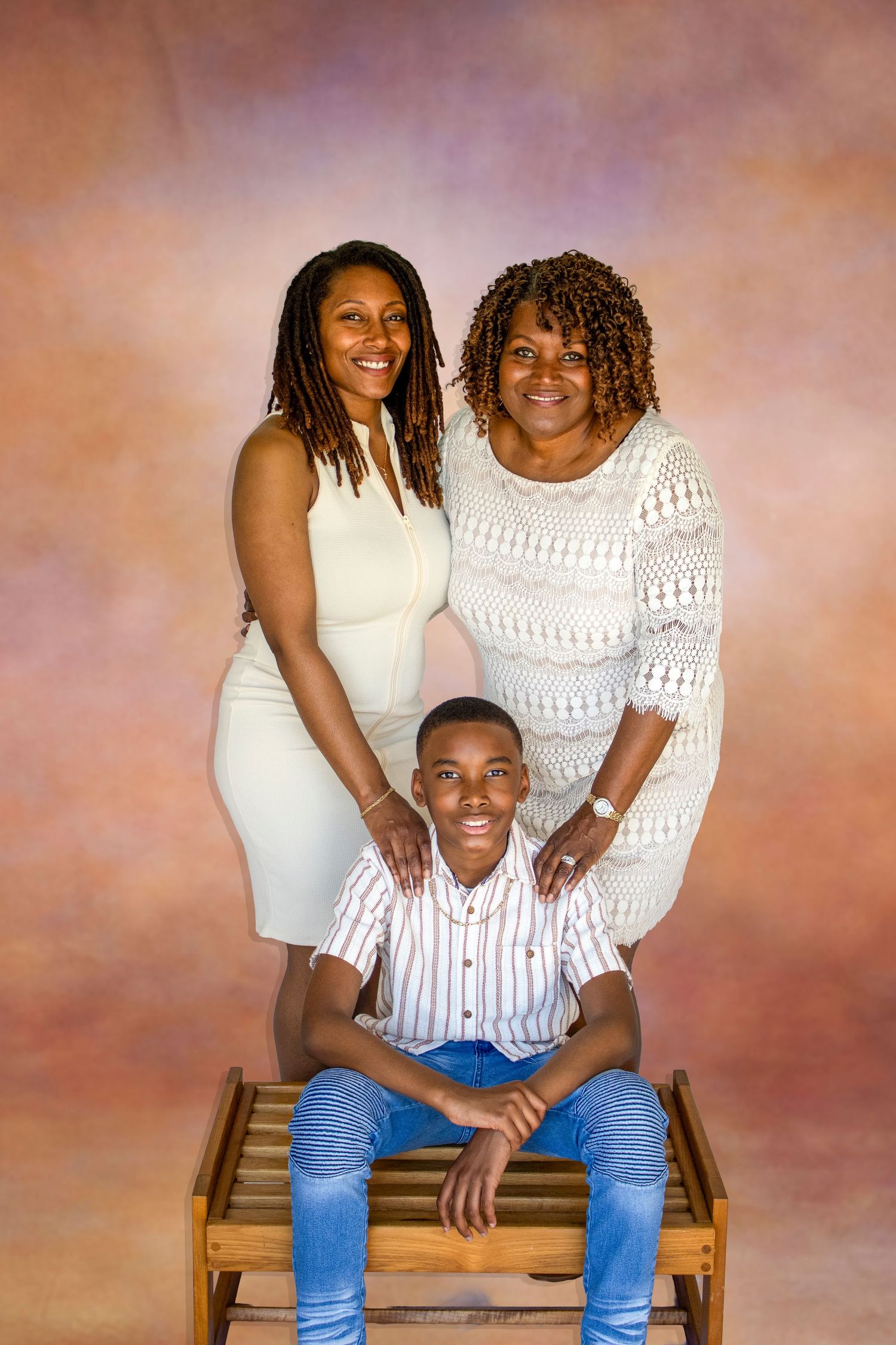 A family portrait of two women and a young man seated on a wooden bench against a painted studio background.
