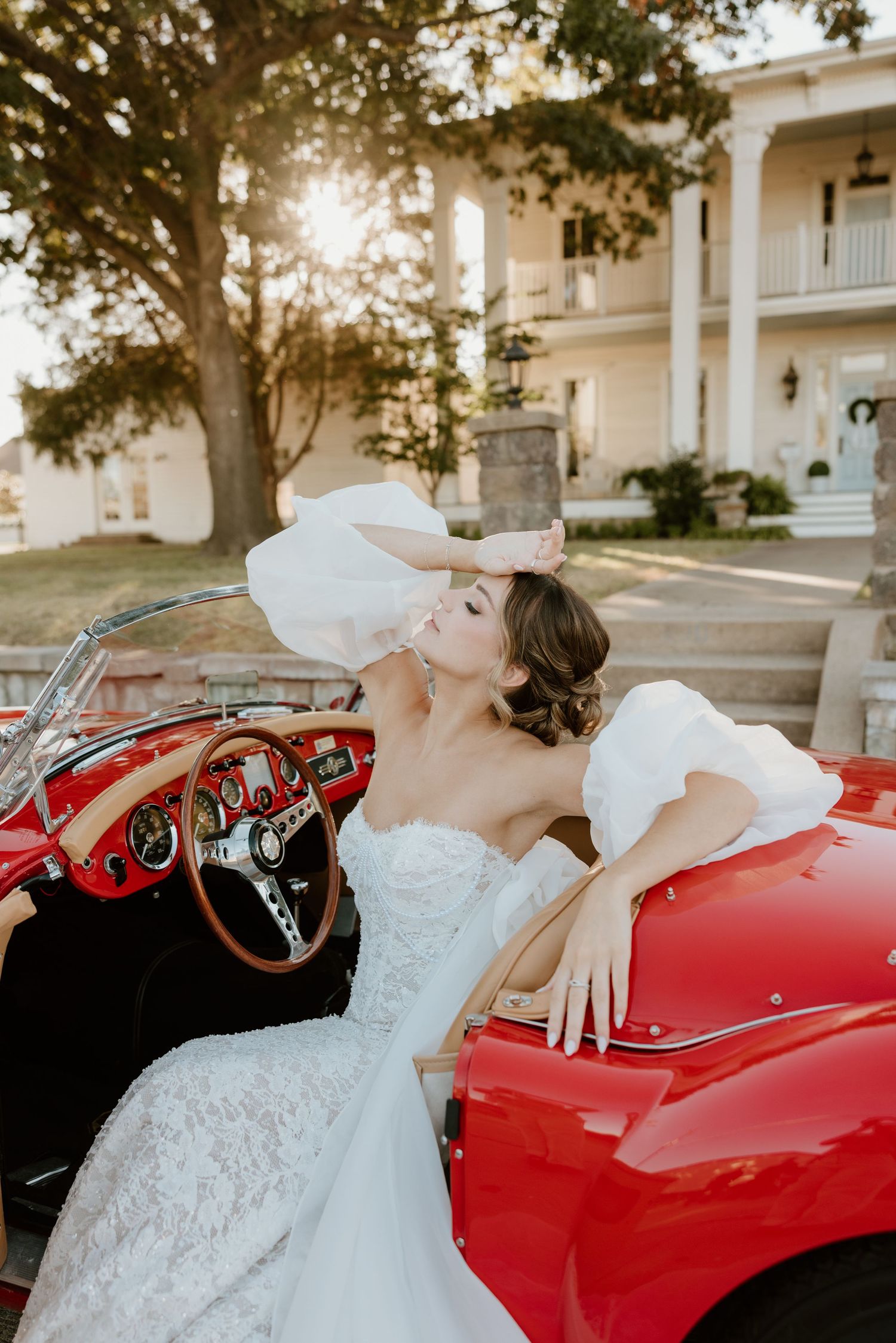 A classic red vintage convertible car is showcased at sunset with someone in a white wedding dress lounging on it.
