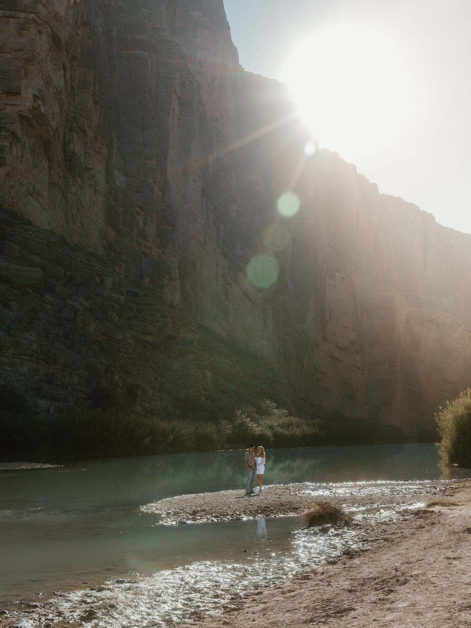 A distant couple stands on a rocky riverbank surrounded by towering canyon walls.