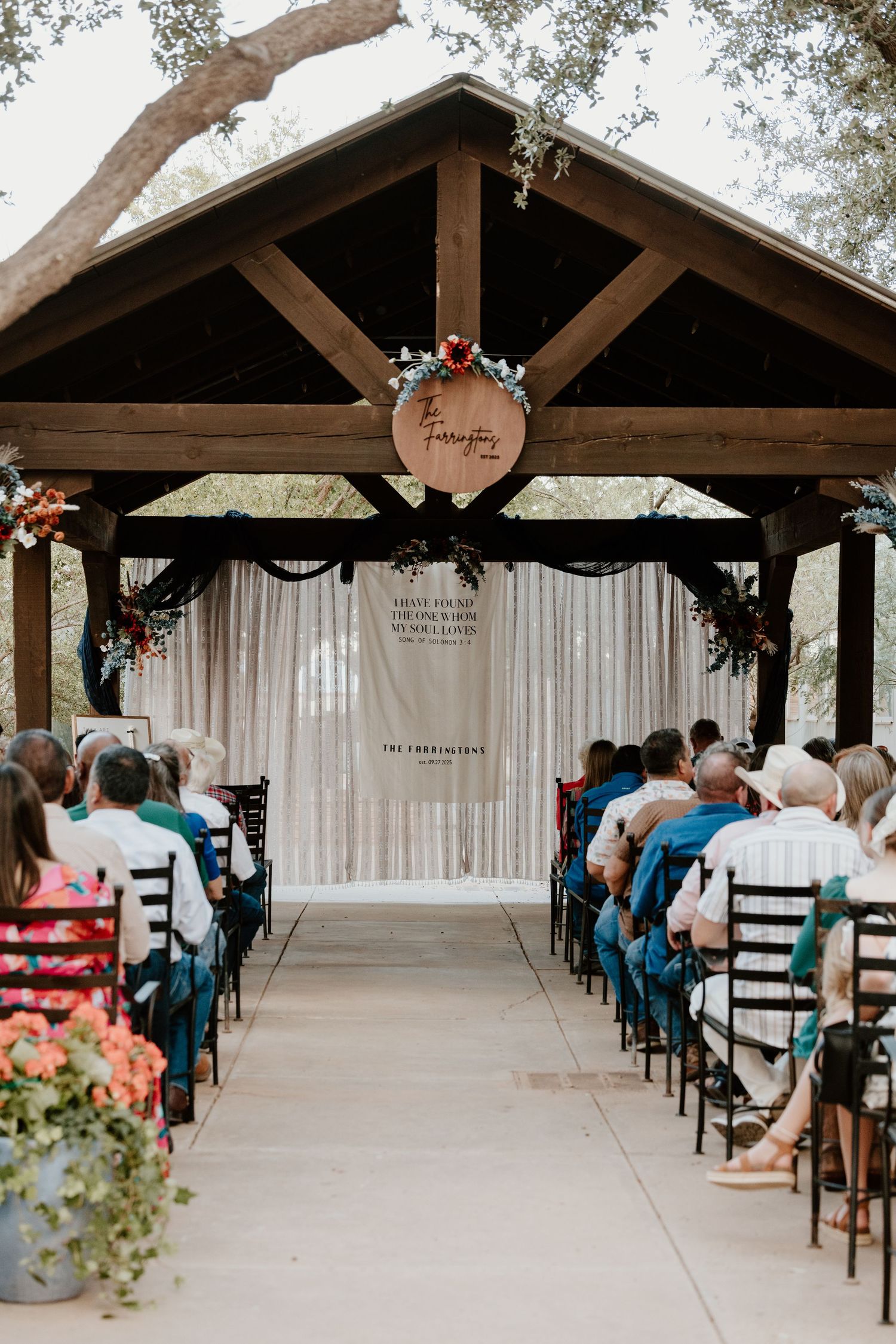A wedding ceremony in progress under a decorated wooden pavilion with white draping.