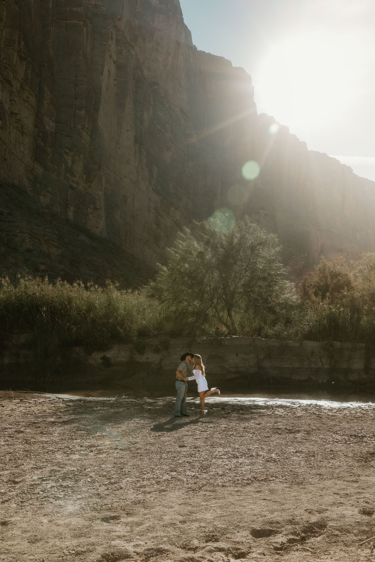 A couple embraces on a rocky riverbank with dramatic canyon walls and sunlight behind them.