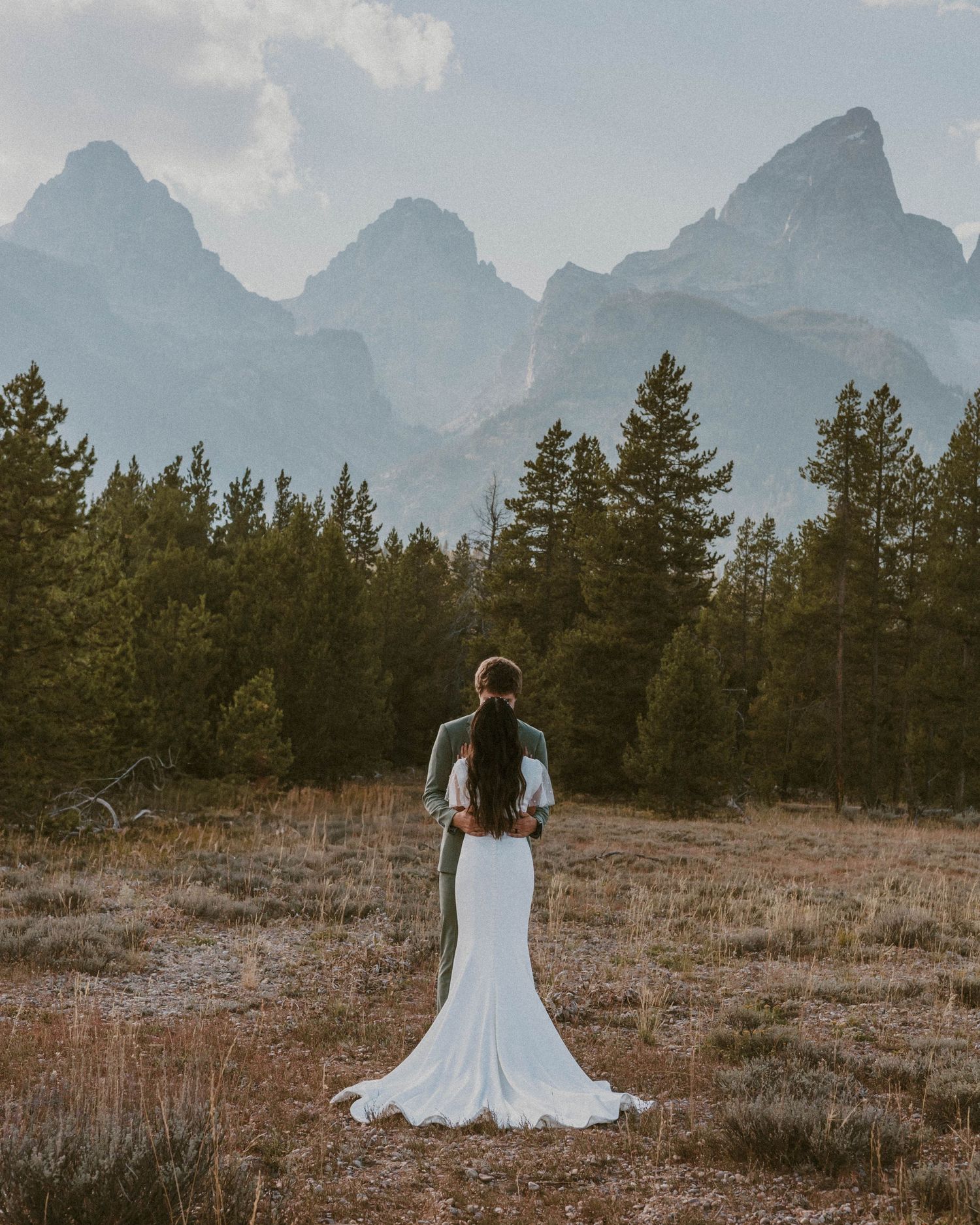 A bride in a flowing white dress stands alone facing mountain peaks in a scenic wilderness setting.