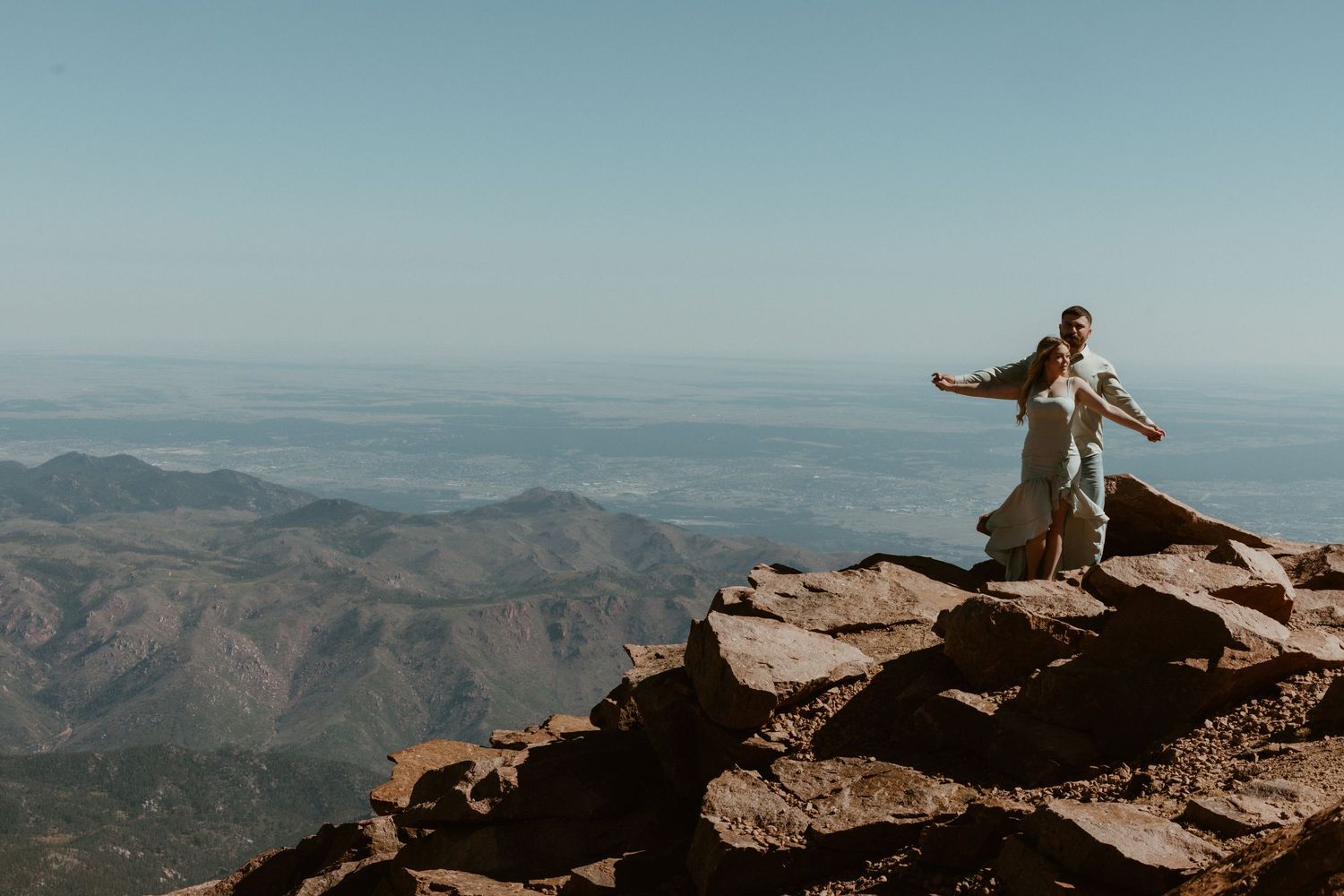 A person stands with arms outstretched on rocky mountain peak overlooking vast mountain ranges.