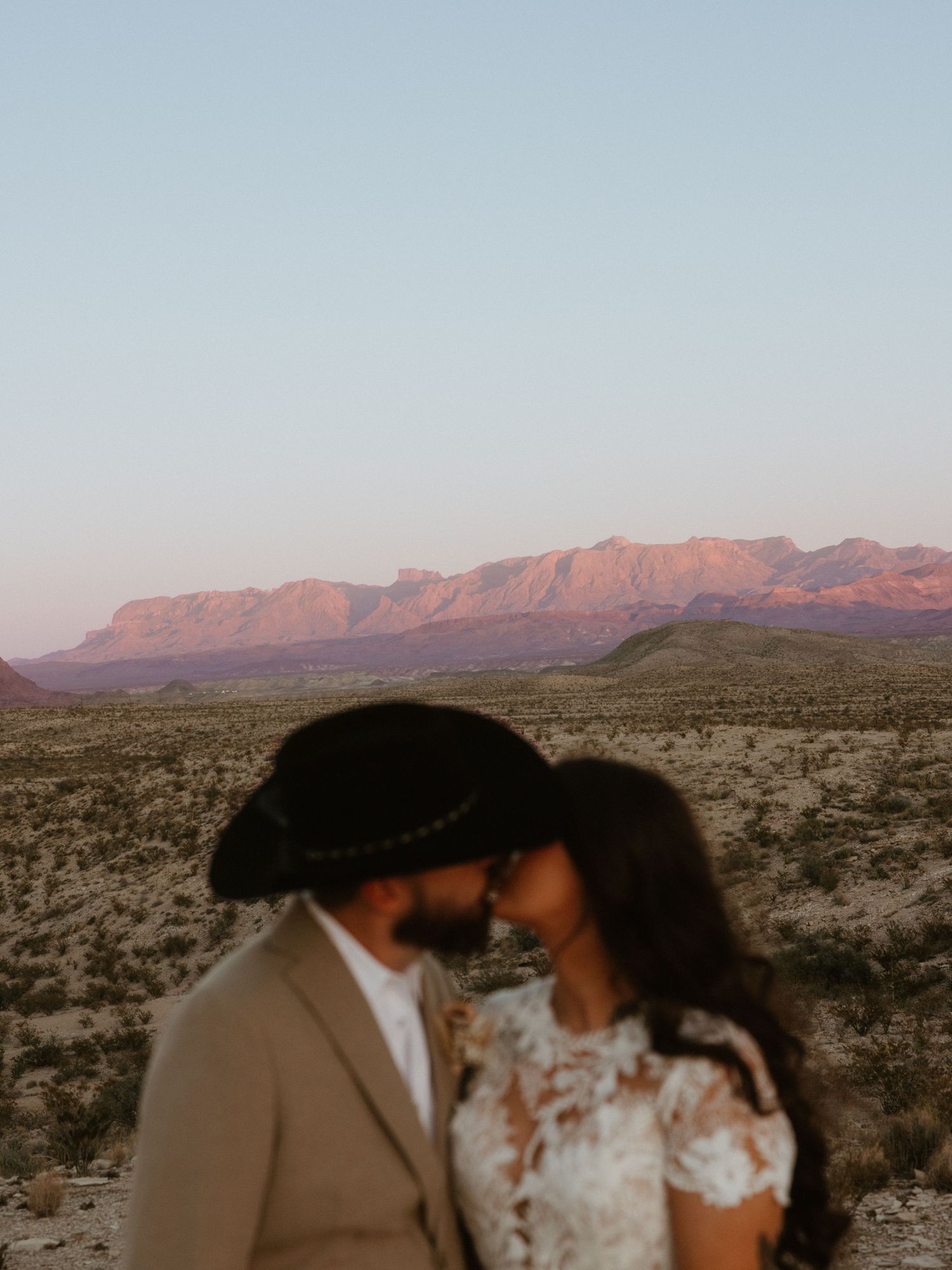 A romantic wedding photoshoot in a desert landscape with mountains glowing pink at sunset.