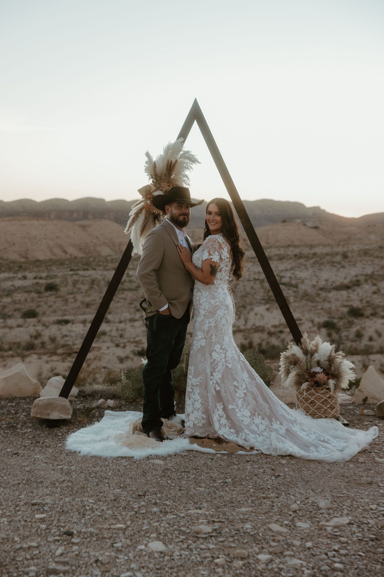 Wedding couple poses under a wooden triangle arch in a desert setting at sunset.