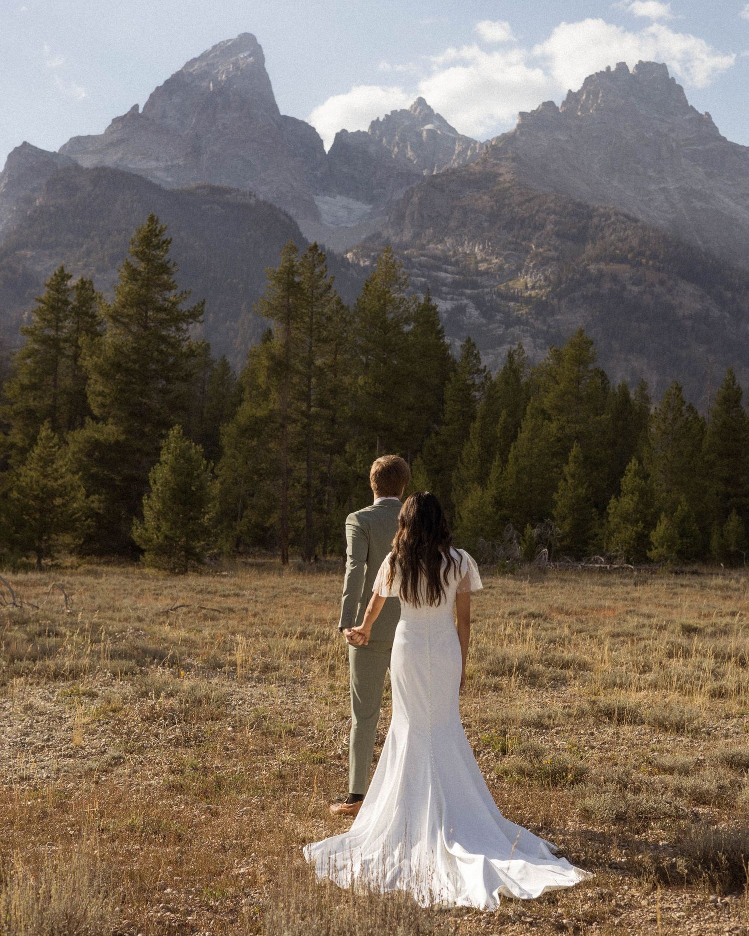Wedding couple walking through mountain meadow with dramatic Teton peaks in background.