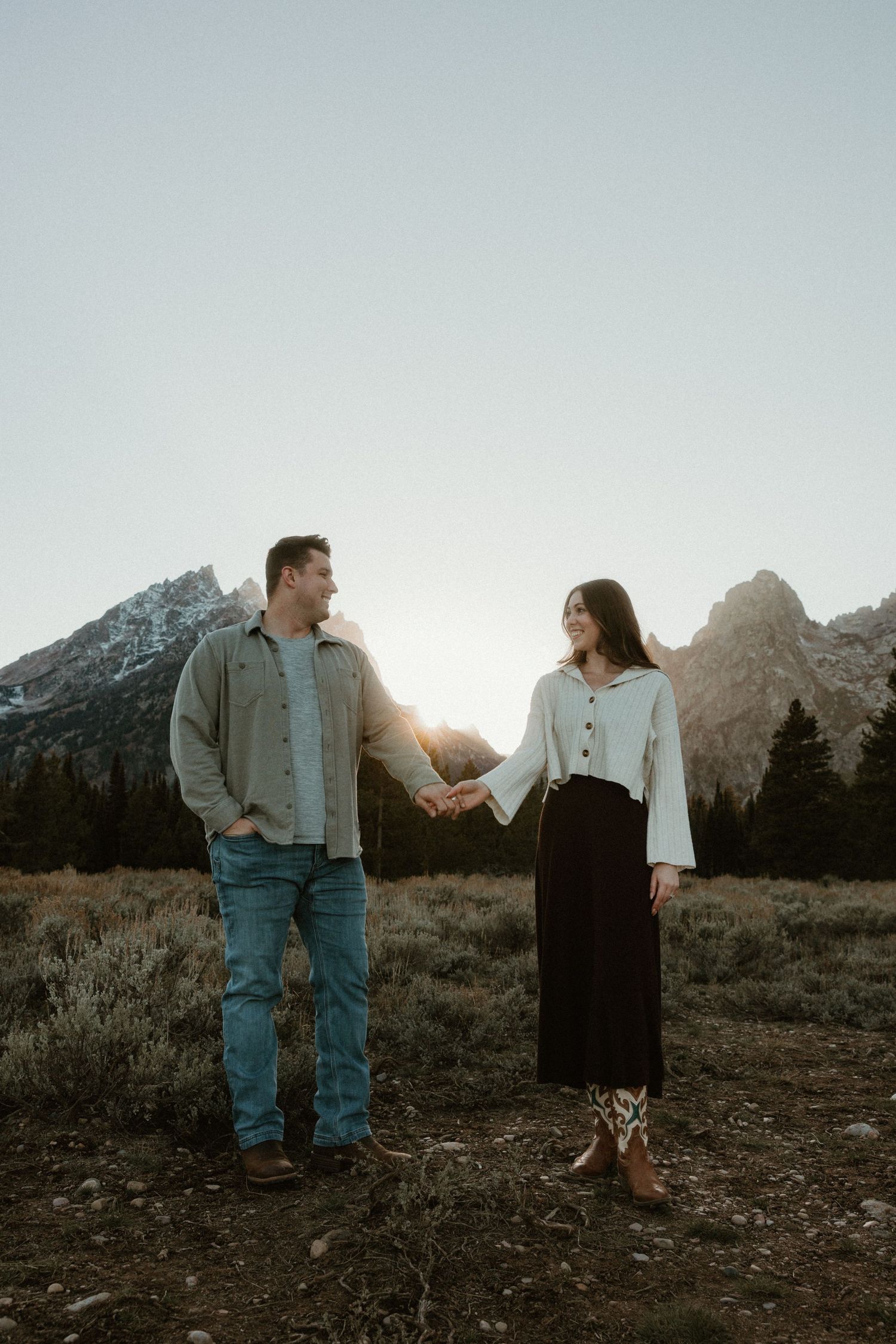 Romantic couple stands together against a backdrop of majestic mountain peaks at sunset in a rustic outdoor setting.