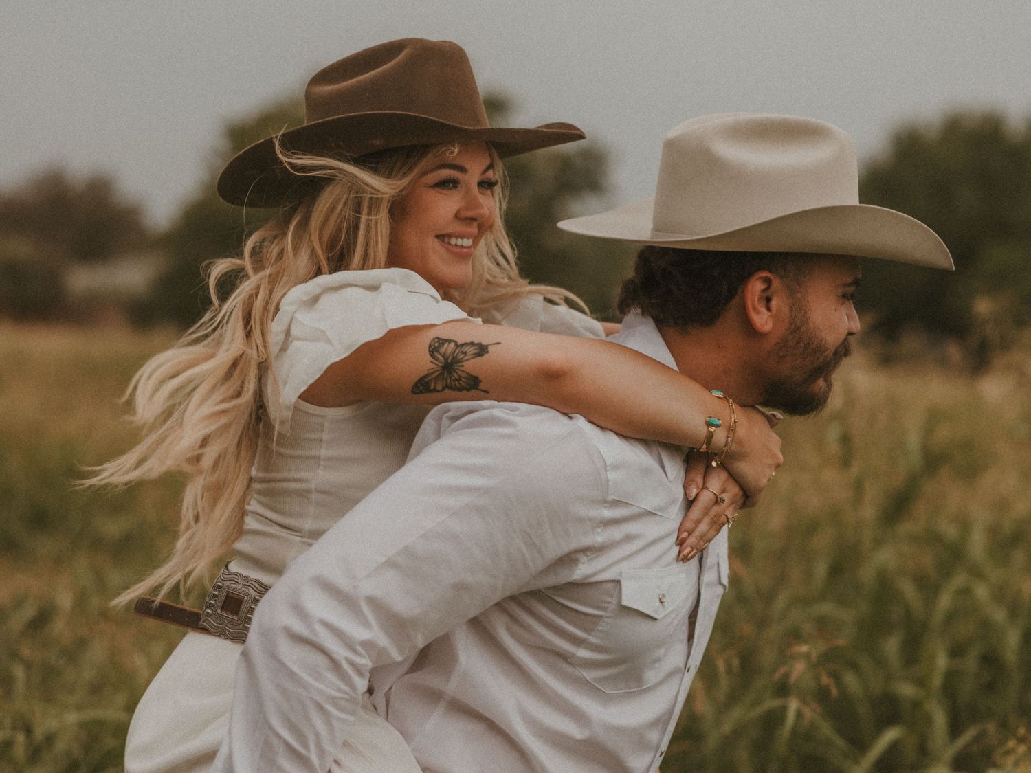 Playful couple in white clothing and cowboy hats embracing in a meadow.
